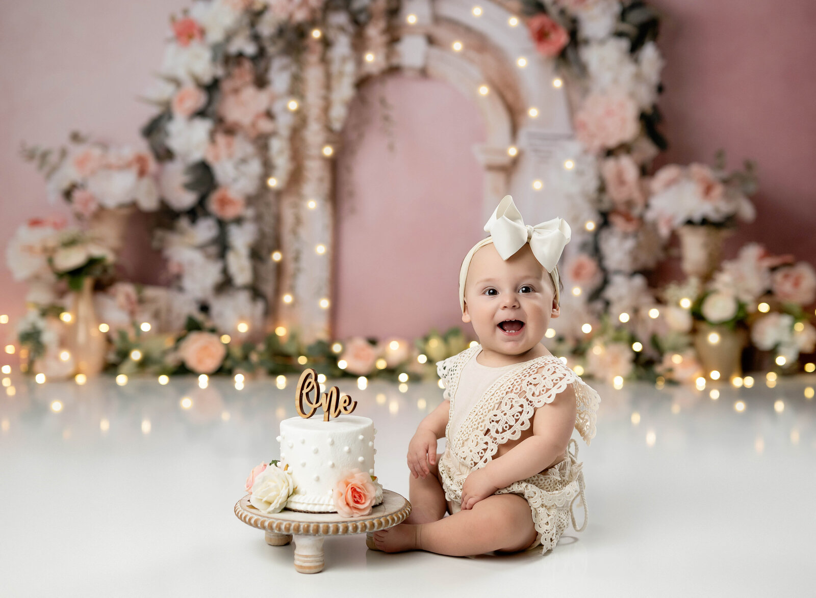 Baby girl in cream lace romper, sitting by birthday cake and smiling. Photographed by Jenni Nikki, Atlanta photographer.