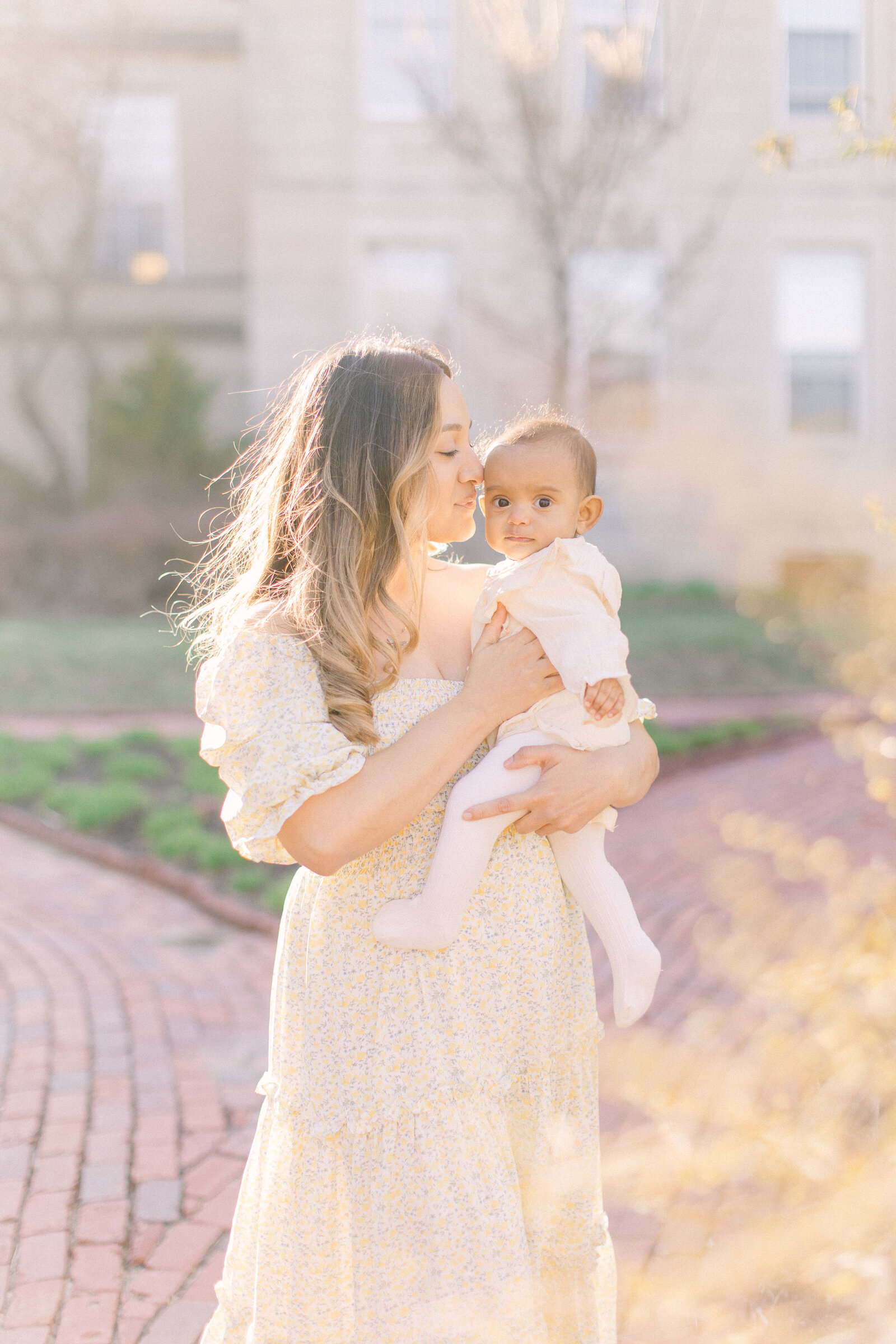 A young mother is holding her baby girl outside standing on a brick pathway in front of some light yellow flowers during photo session with Boston newborn photographer Corinne Isabelle