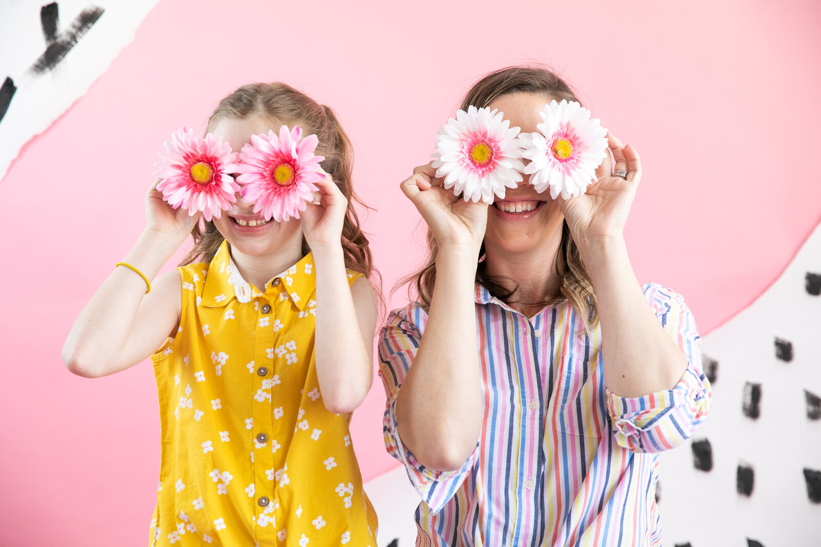mom and daughter in front of pink backdrop holding pink and white daisies in front of their eyes