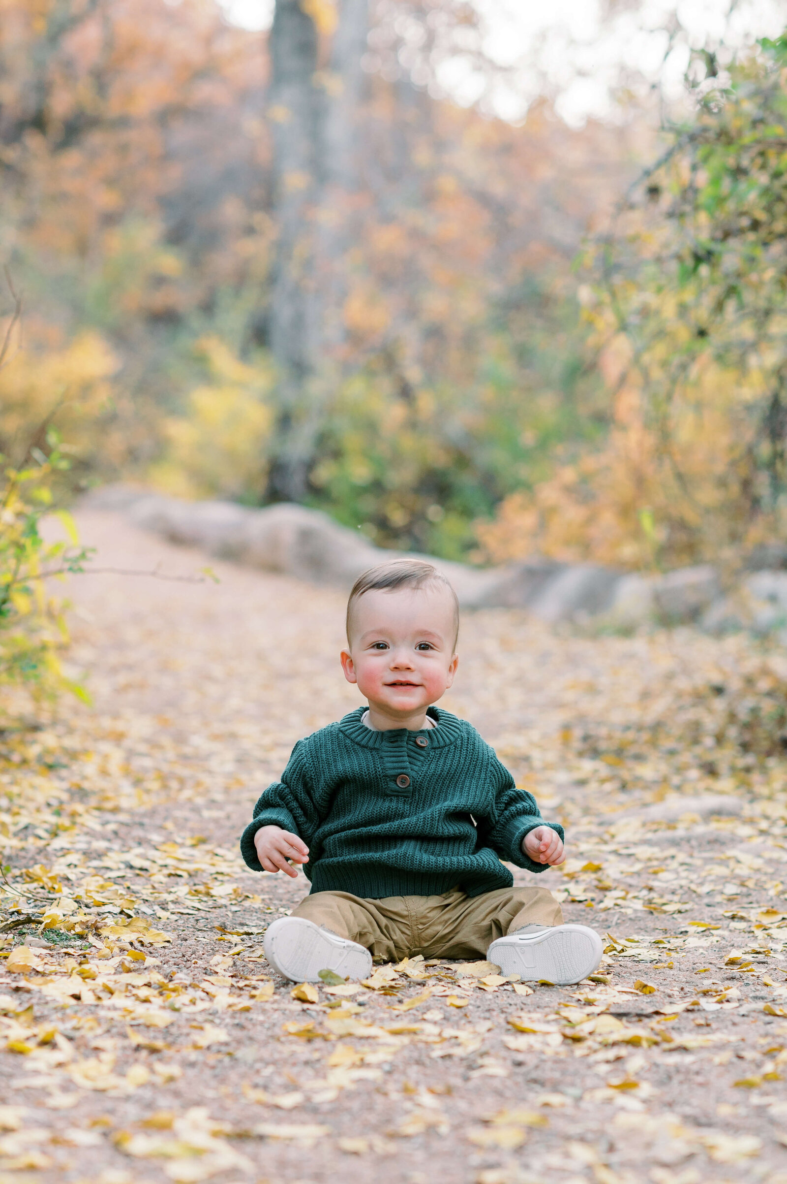 A young baby wearing a green sweater and tan pants sits on a path that is covered with fall leaves