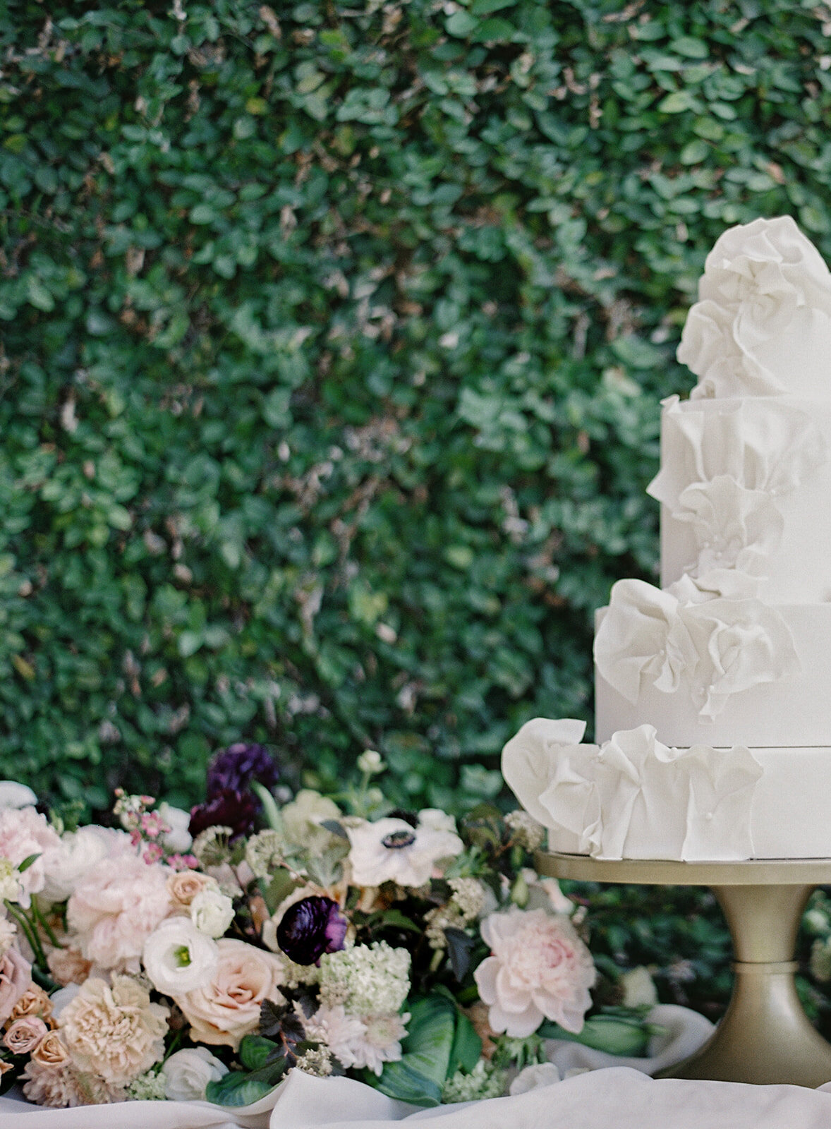 White wedding cake on gold pedestal with bold florals against an ivy wall. Cake is three tiers with icing creating ruffles coming off of all three tiers. Photographed by wedding photographers in Charleston Amy Mulder Photography.