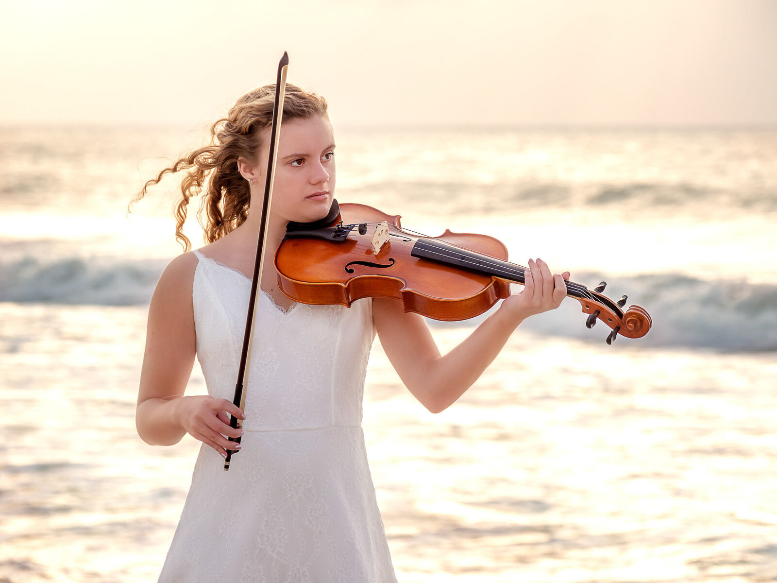 A high school senior in a white dress playing the violin on the beach with ocean waves in the background in North Carolina. This artistic portrait captures the harmony of music and nature, ideal for those seeking unique senior photography in Wilmington, NC.