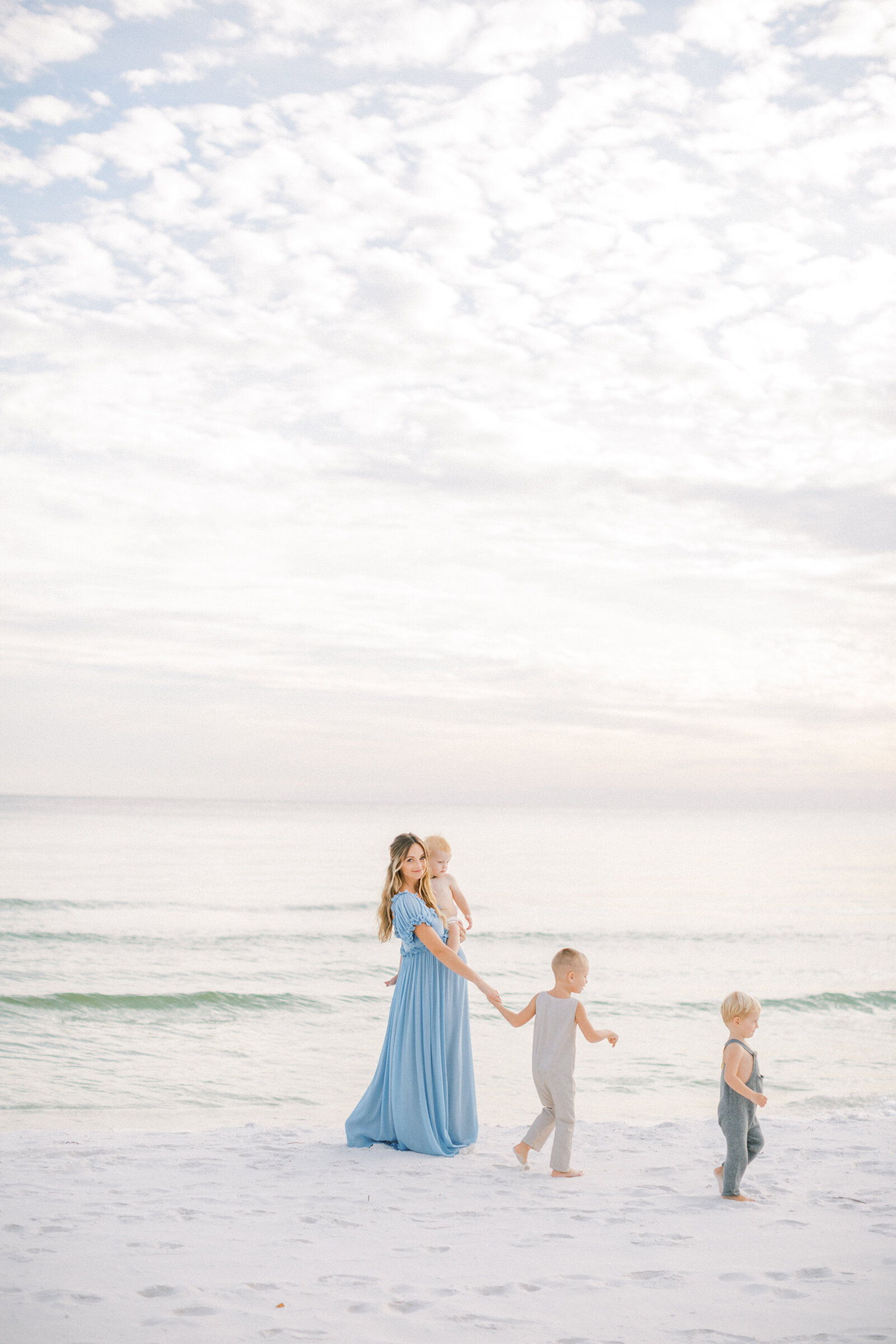 An expecting mother wearing a blue gown walks on the beach with her three sons