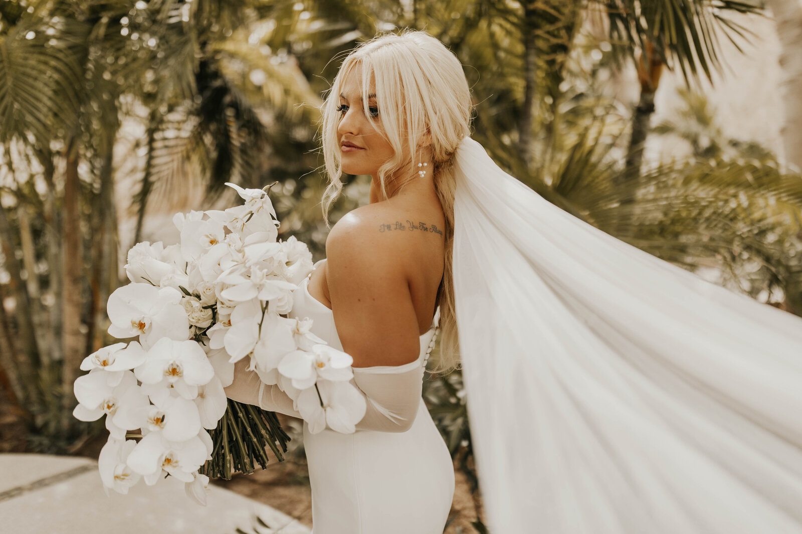 Blonde bride holding florals with long veil in front of palm trees