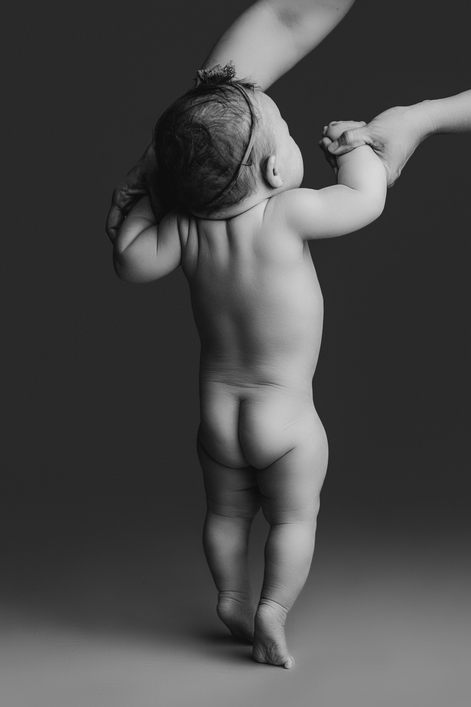 Minimalist black and white milestone image of a baby girl standing with tushy showing, supported by a parent’s hands during a milestone photography session.