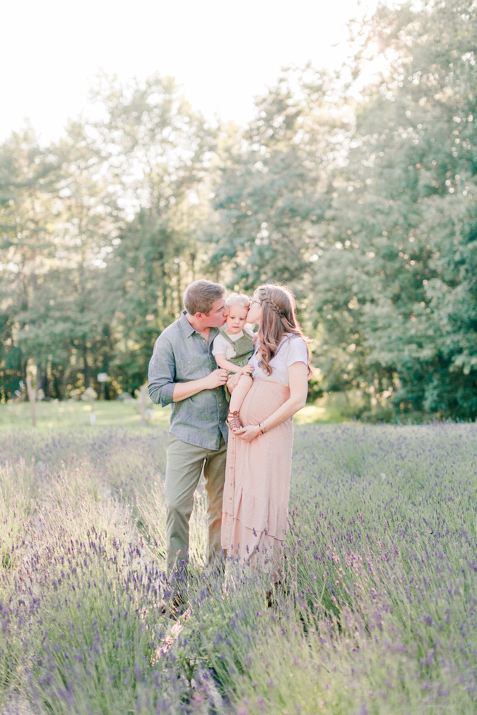 Pregnant mom and dad hold their three year old daughter and kiss each side of her head in a field of lavender by NH newborn photographer Fieldstone Studio.