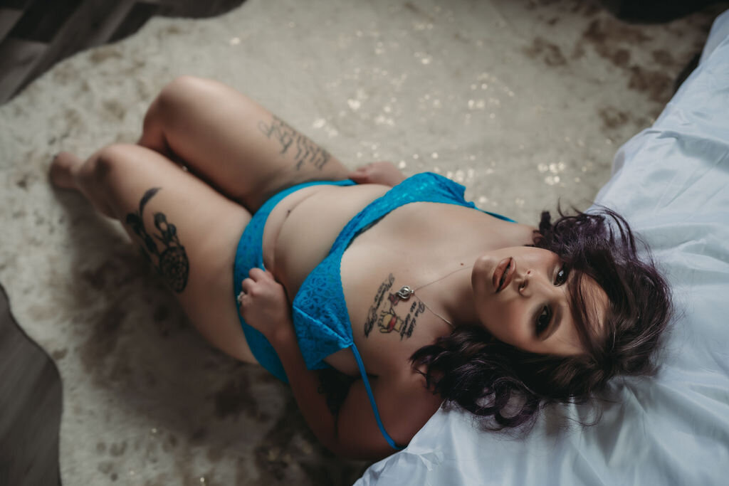 plus size woman in blue lingerie leaning against bed with white sheets on ground boudoir