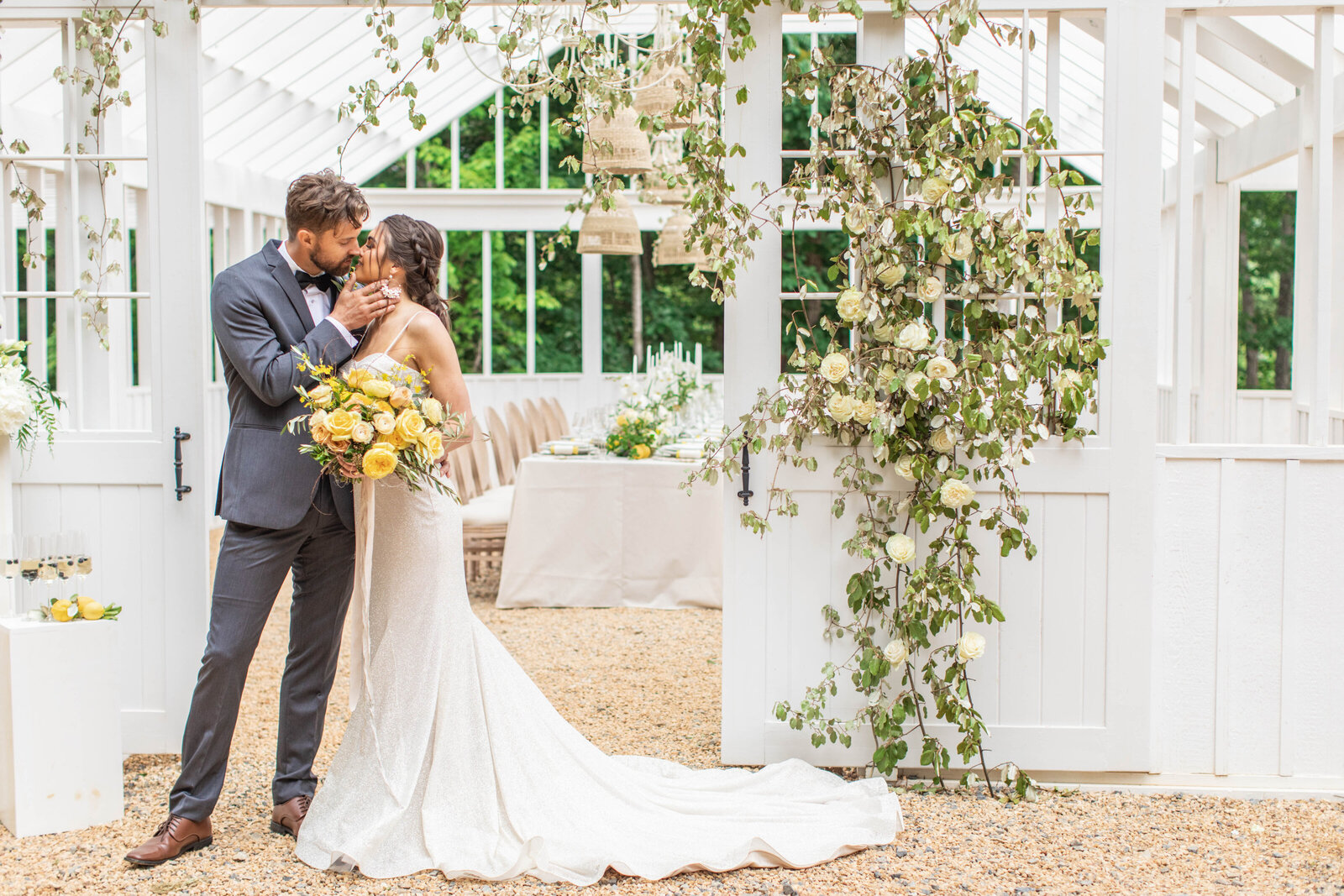 Classy couple kiss on their wedding day surrounded by greenery and holding a big yellow  flower bouquet