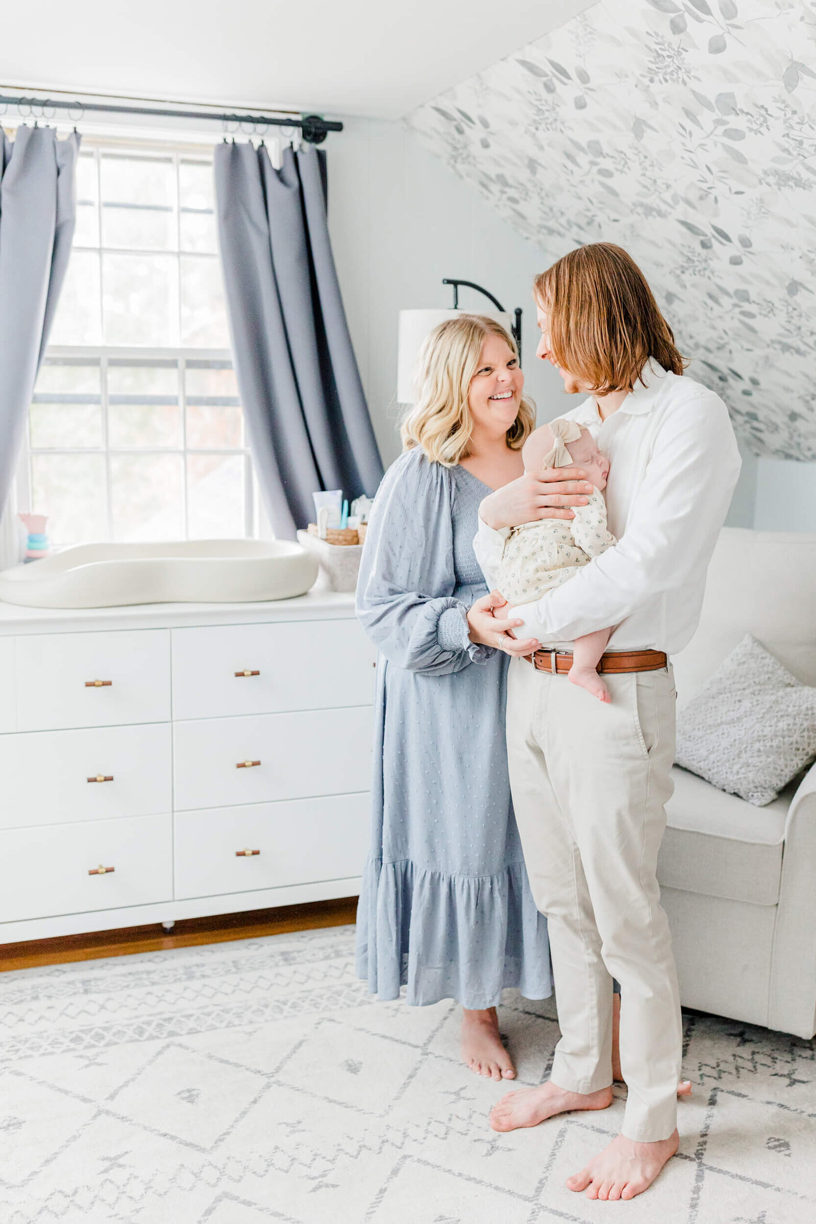 Mom and dad stand smiling at each other while dad holds their newborn in the nursery