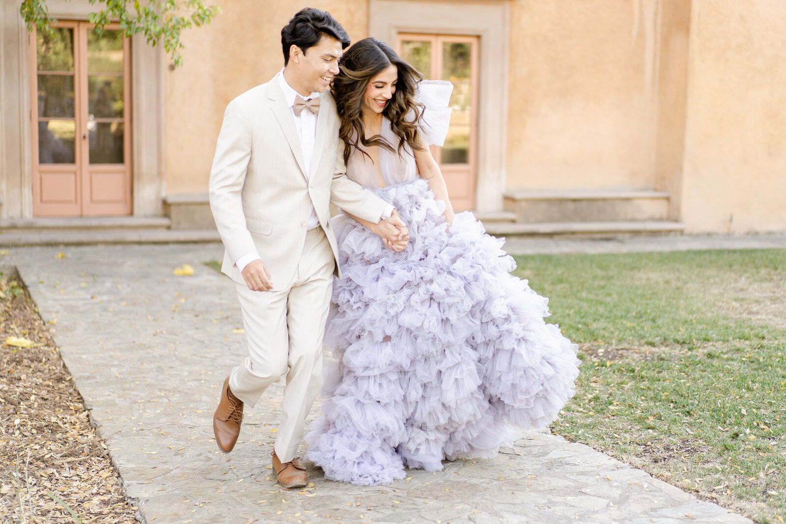 Portrait of bride and groom in a lavender wedding gown and cream suit in front of a Spanish-style venue