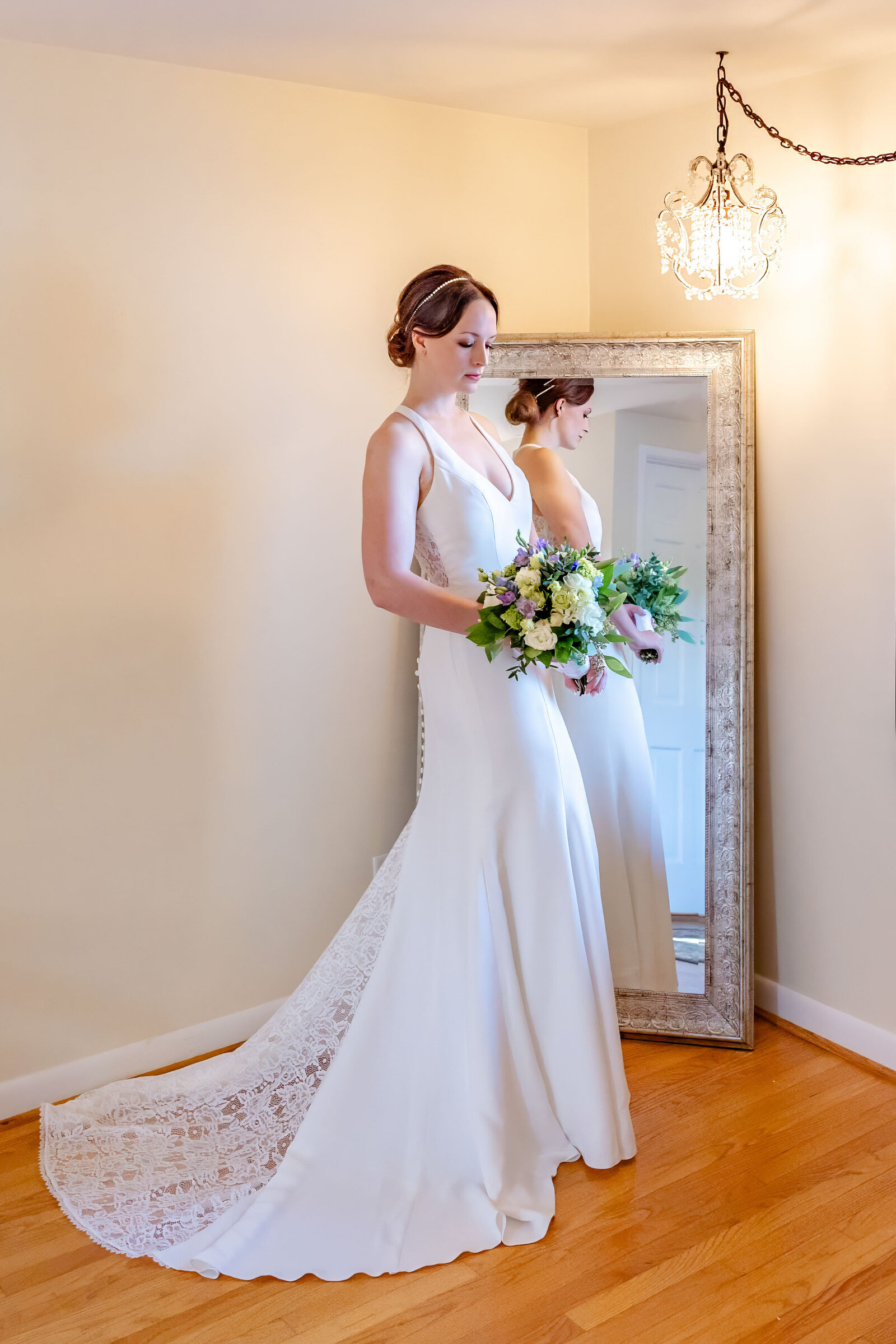 A bride in a sleek, fitted gown holds a vibrant bouquet while admiring her reflection in a vintage mirror, captured in a bridal suite during an Asheville, NC wedding, showcasing timeless elegance and natural beauty.
