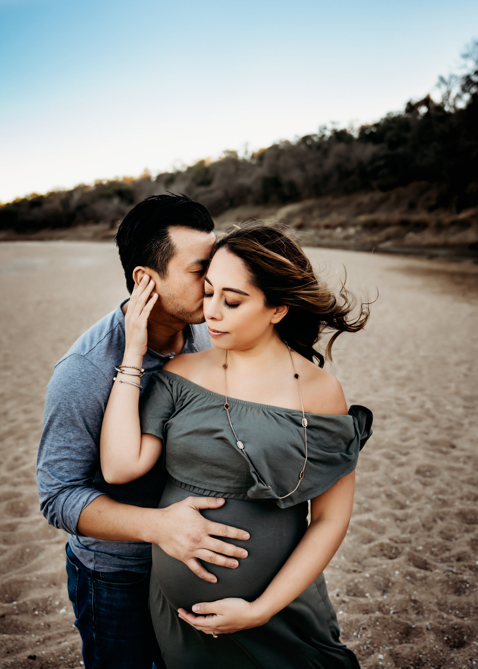 Maternity Photographer, a husband kisses his pregnant wife as he holds her belly on the beach