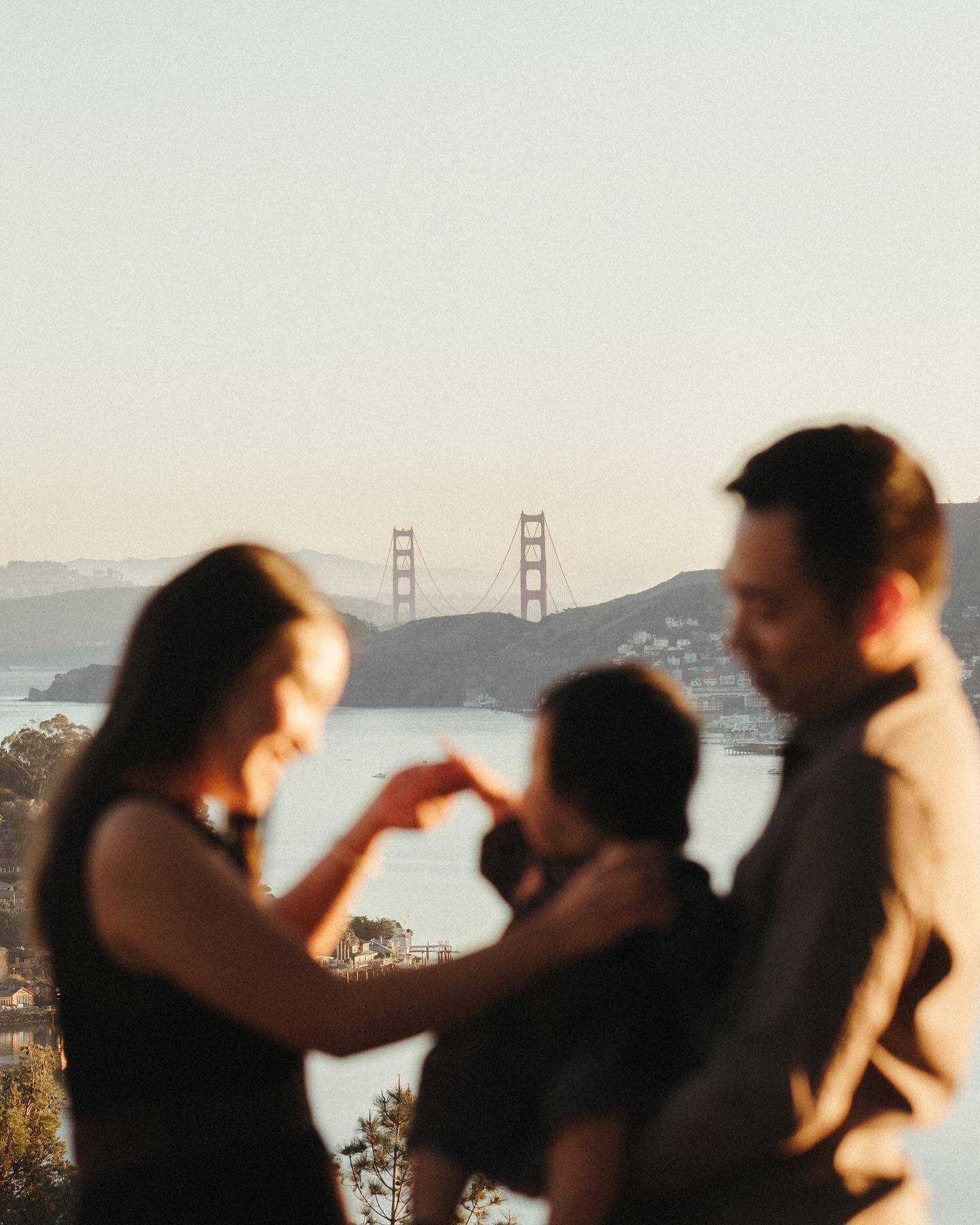 A family of three, with a blurred foreground of a couple and a child, sharing a cheerful moment during their family photoshoot against a backdrop of the golden gate bridge and blue sky in San Francisco.