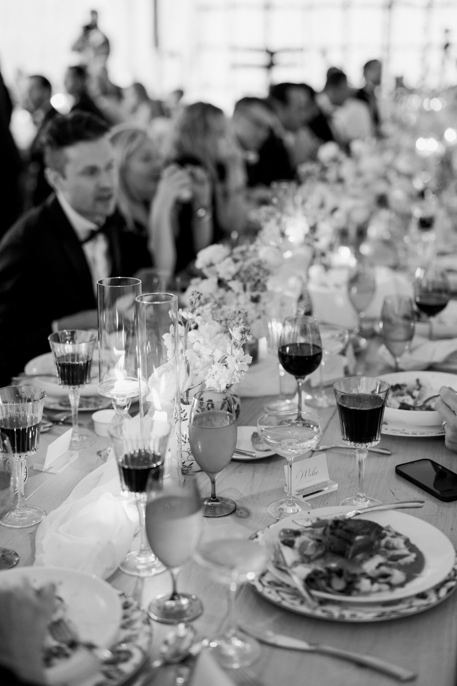 Black and white photo of a long wedding reception table as people eat dinner.