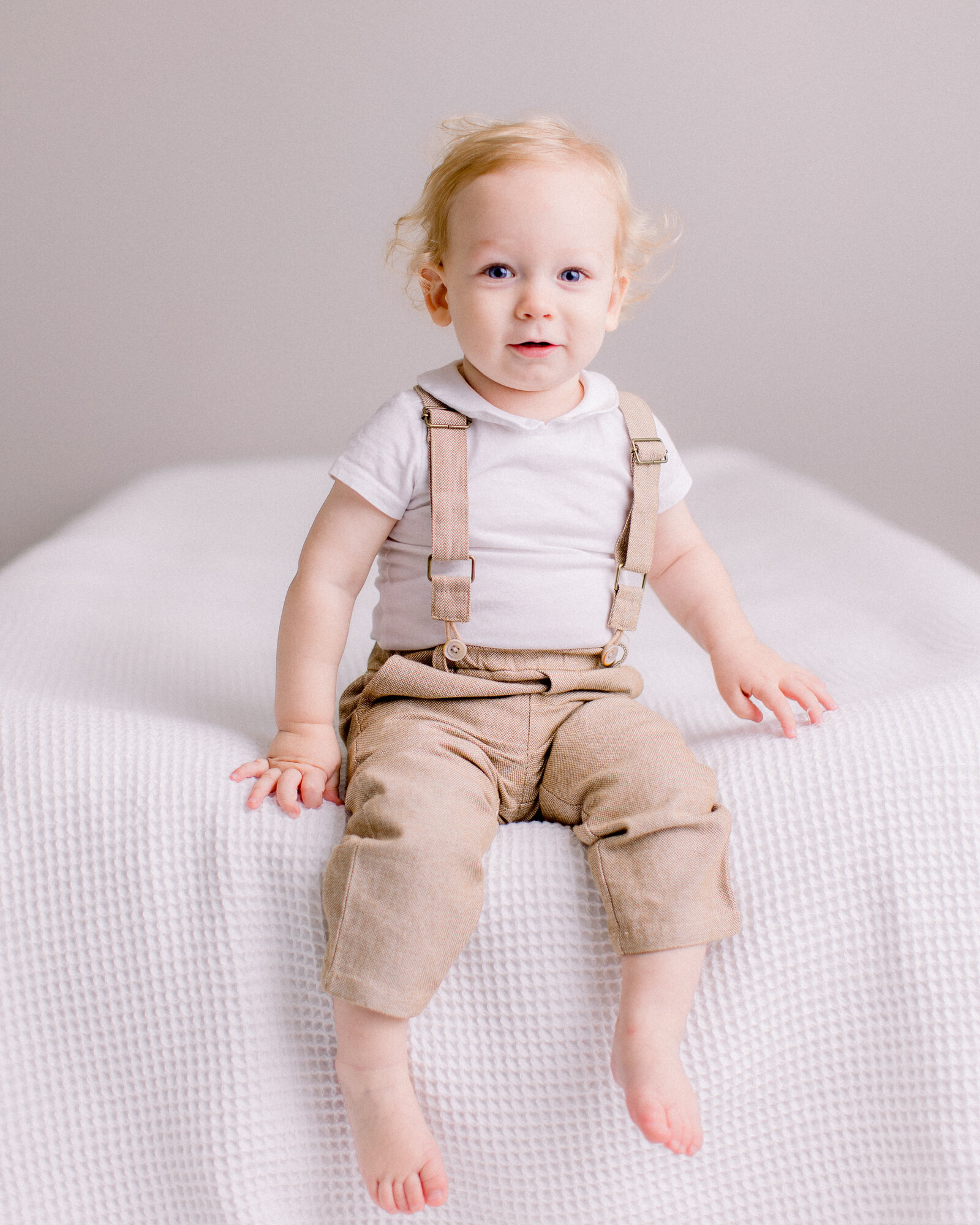Two year old sitting up on the edge of a bed smiling with his feet dangling by NH newborn photographer Fieldstone Studio.