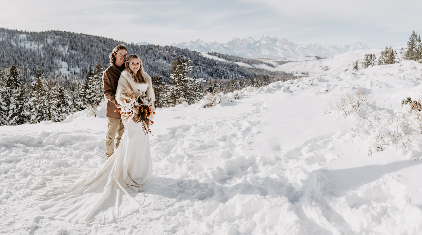 grand teton winter wedding with bride and groom on an outlook in the Wedding Tree with snow surrounding the couple with a forest of trees in the distance covered in snow captured by photographers jackson hole