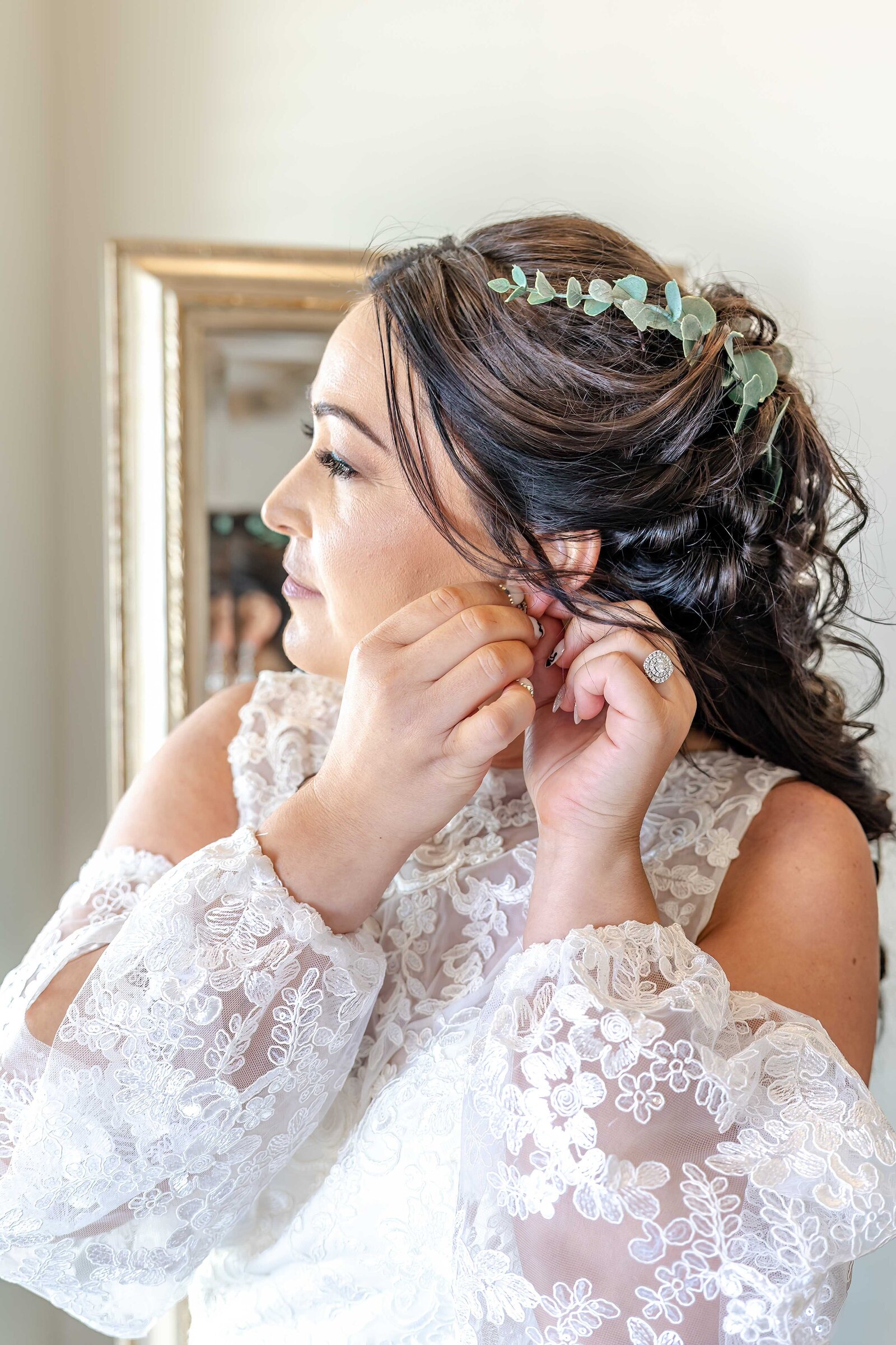 Bride putting on earrings while preparing for her wedding, showcasing intricate details of her lace dress and floral hairpiece in Ocean Isle Beach, NC.