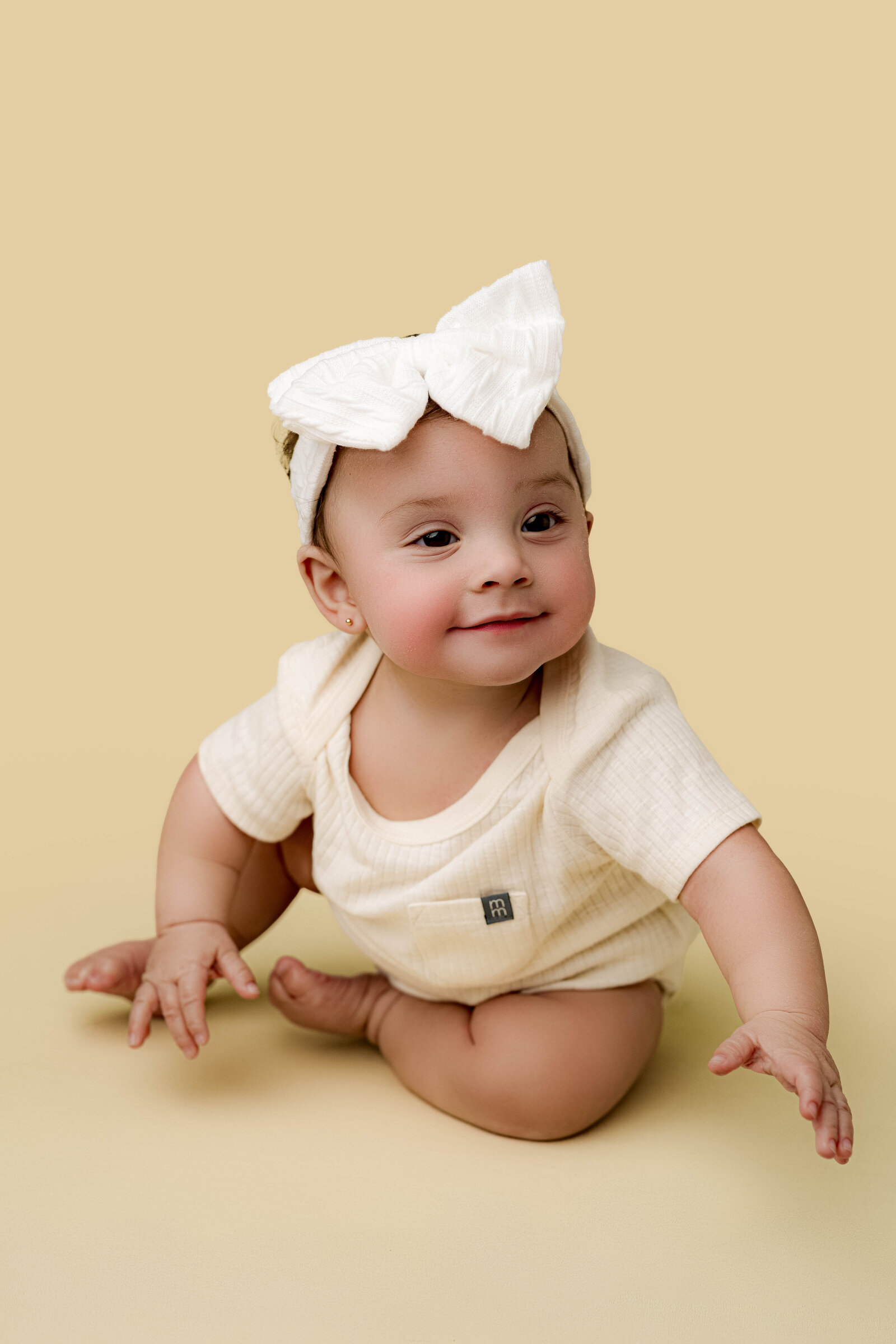 Baby girl wearing a white bow headband and cream onesie, sitting and leaning forward on a soft yellow backdrop in a Charlotte milestone session.