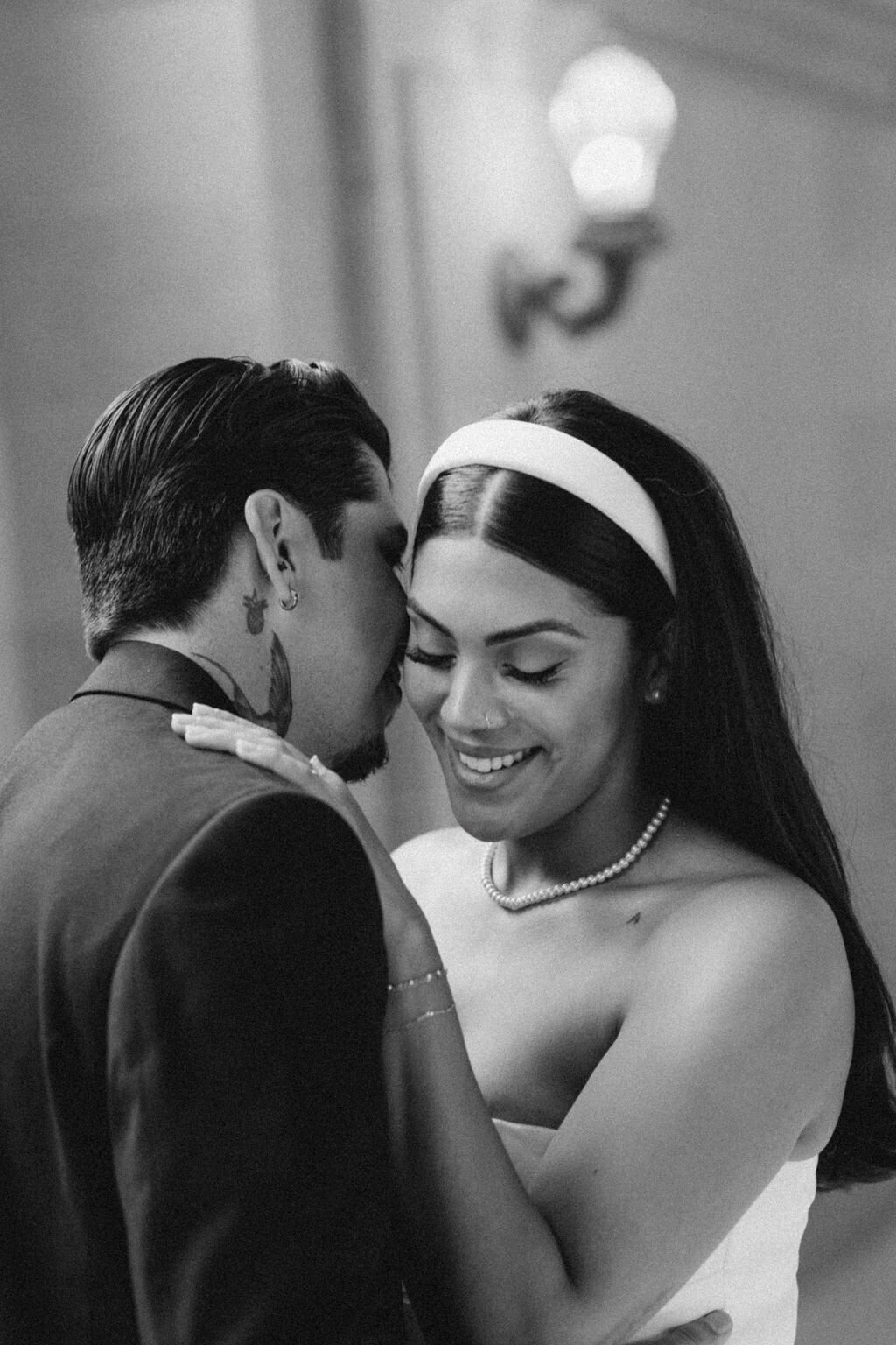 A black and white photo of a groom embracing his bride as she smiles, both dressed in elopement attire with the bride wearing a pearl necklace and headband during their photoshoot in San Francisco