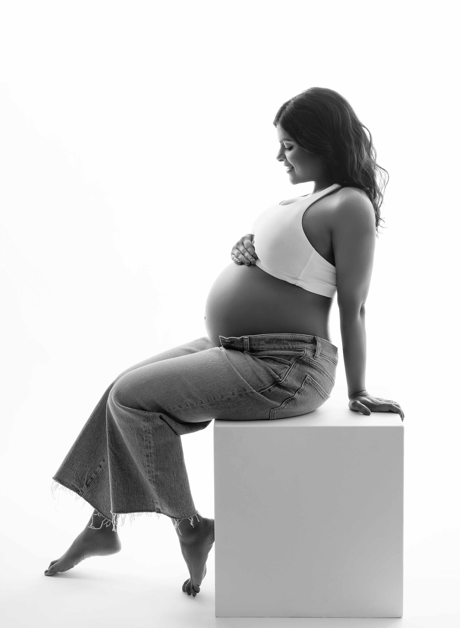 Mom dressed in jeans, sitting on white box and looking at baby bump. Pregnancy photo by Jenni Nikki Photography at Atlanta maternity photo studio.