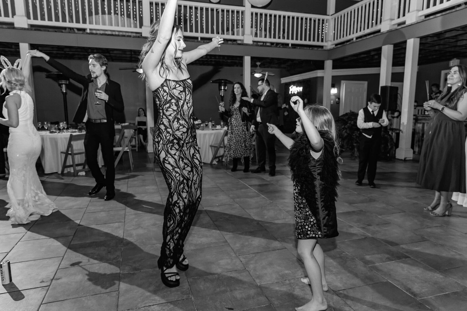 A woman and a young girl dance joyfully together at a formal indoor event, surrounded by other guests who are mingling and watching. The scene is lively and festive, with people dressed in elegant attire.