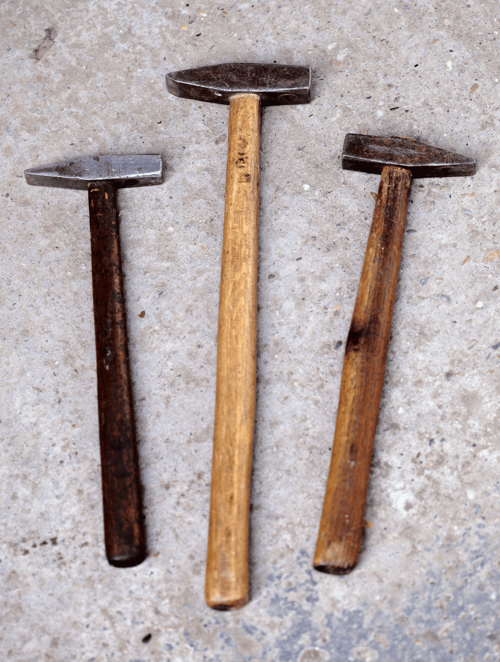 Three tack hammers with worn wooden handles sitting on warm grey concrete