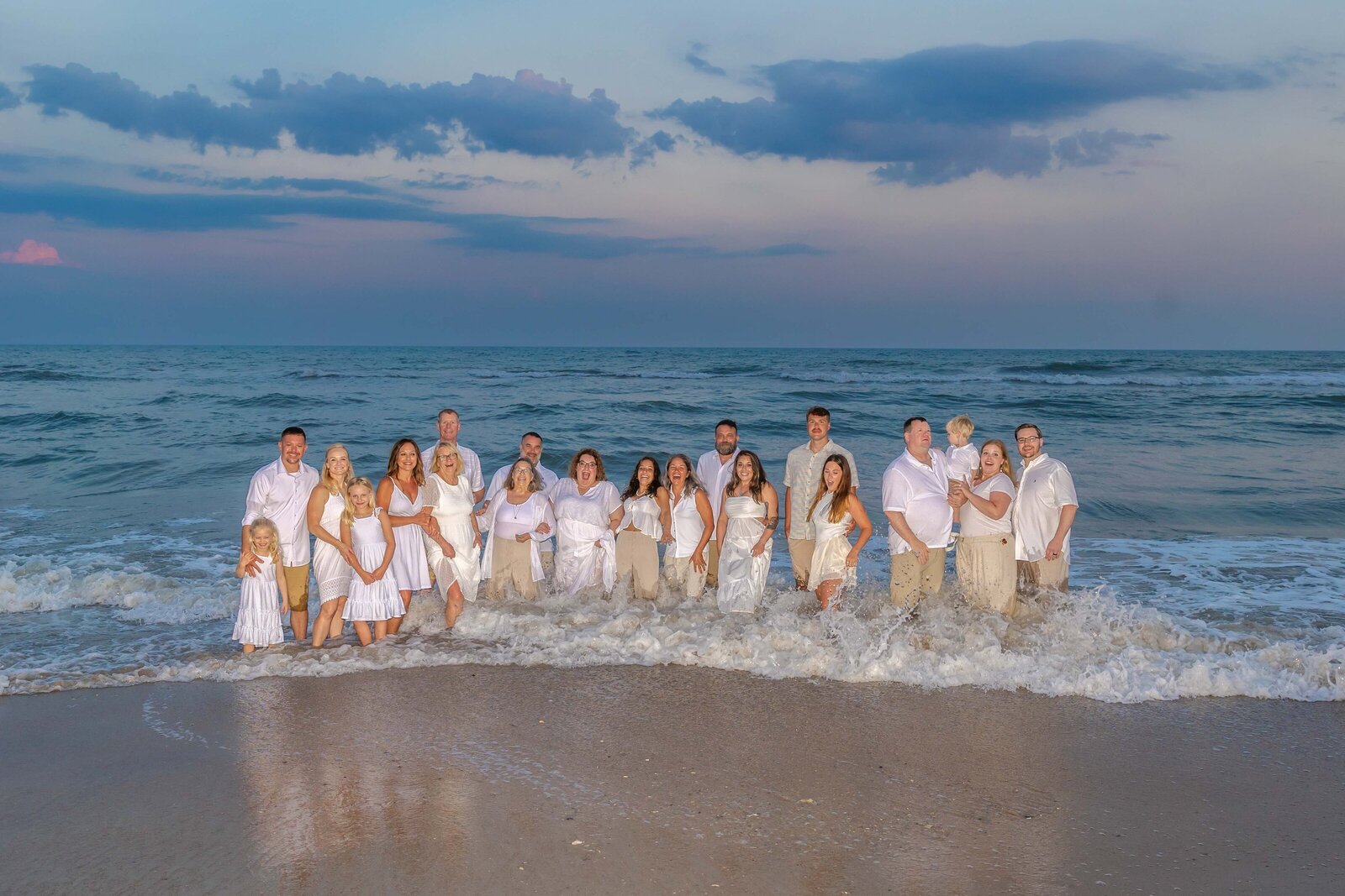 A large family, dressed in white, stands in the gentle ocean waves at sunset. The dramatic sky enhances the scene, ideal for professional family photography in NC, capturing elegant and joyful family moments by the sea.