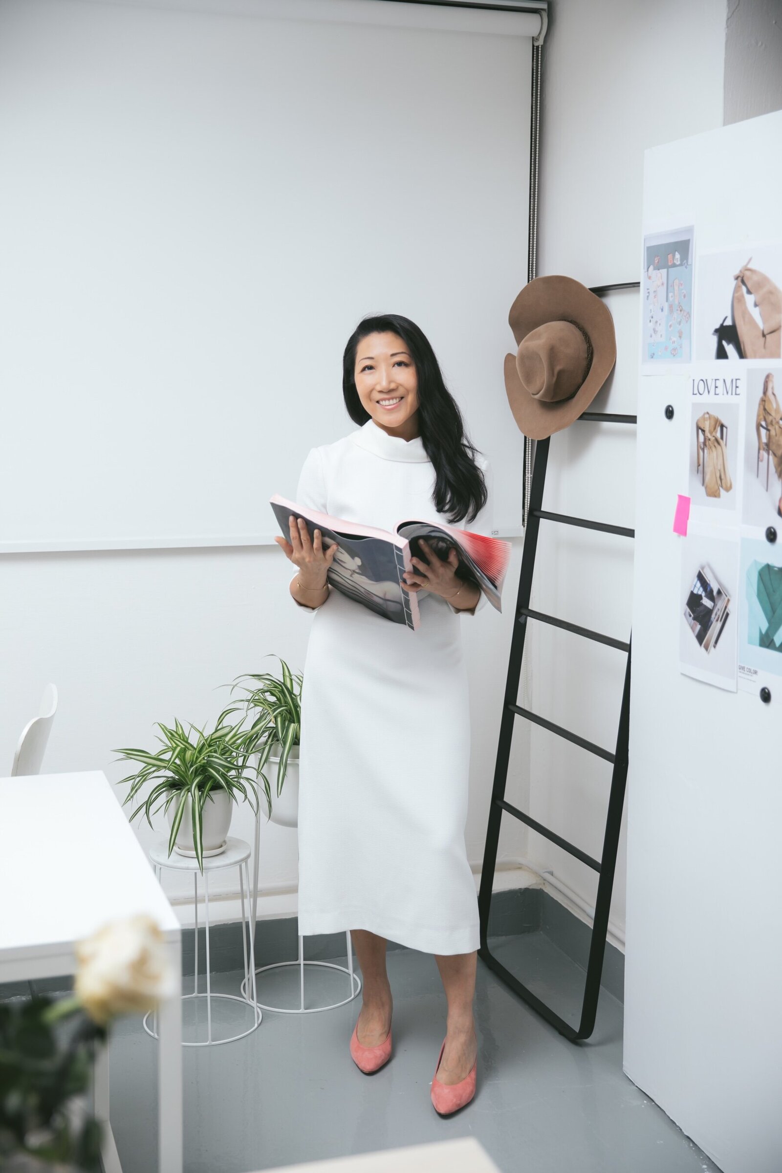 Picture of woman in white dress standing next to moodboard holding a magazine