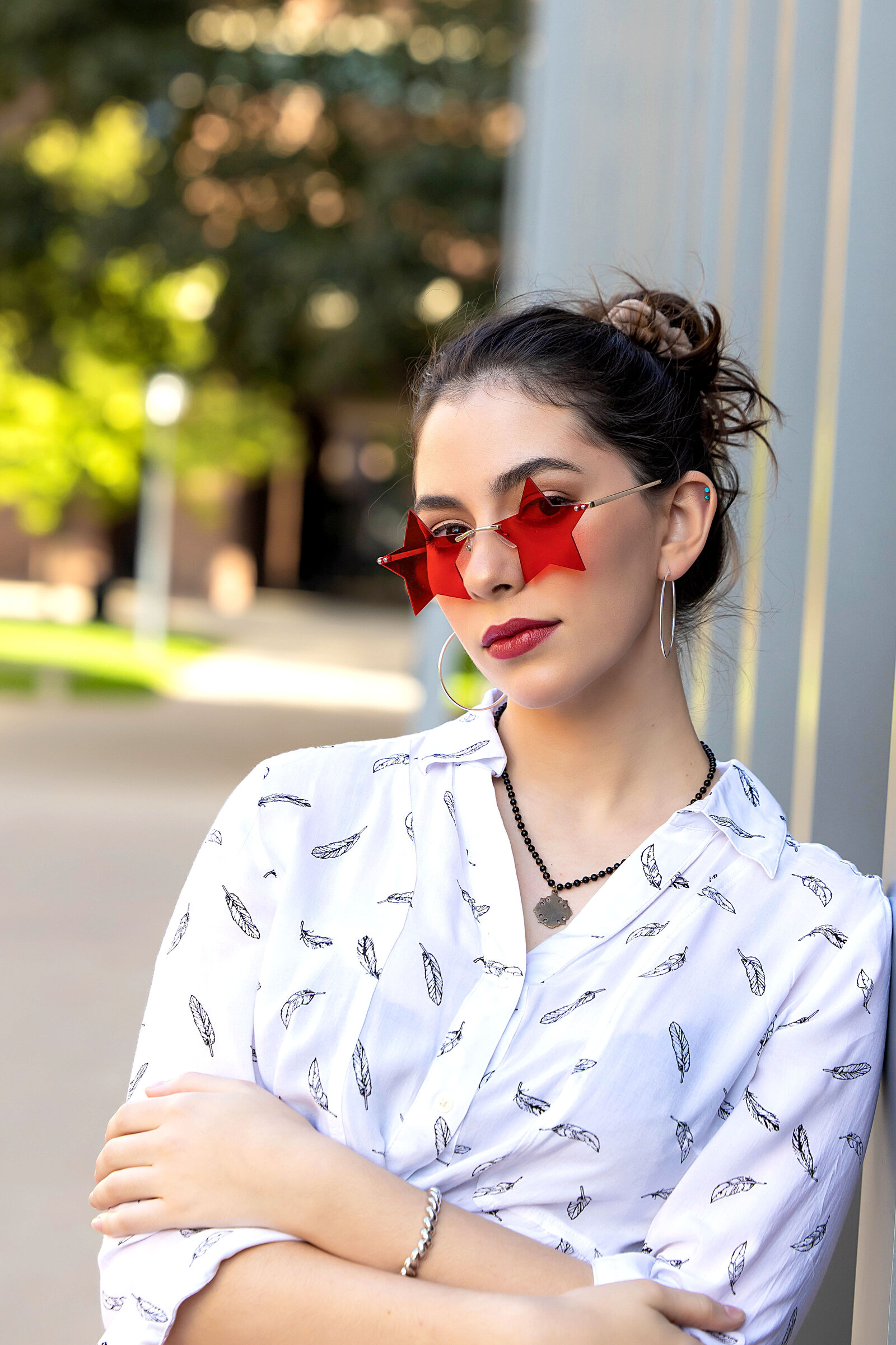 Senior girl in white printed top, red pants, and white boots leaning casually against grey pillars with red star sunglasses