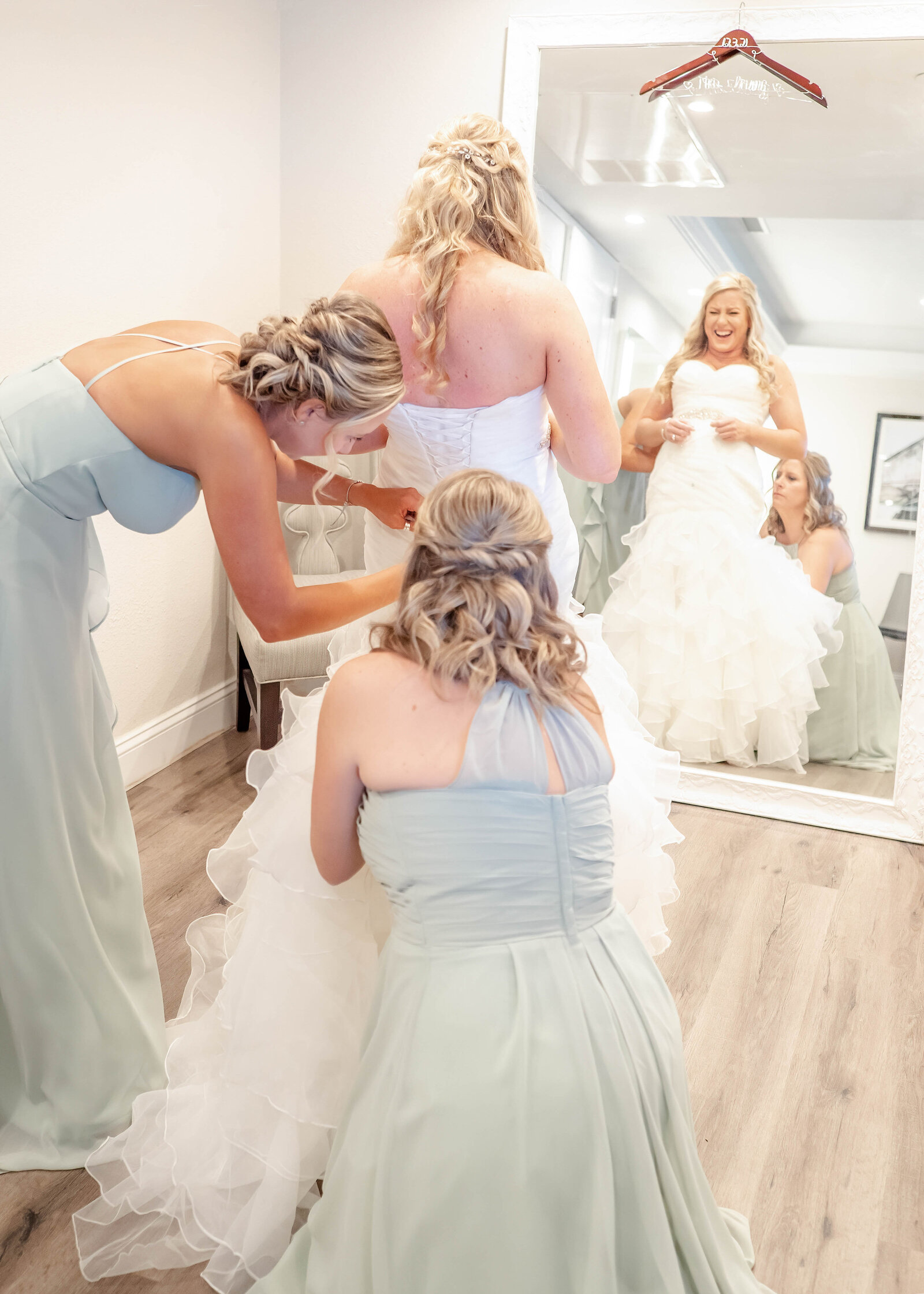 A joyful bride in a ruffled wedding gown is assisted by her bridesmaids in sage green dresses in an elegant bridal suite, during a Wilmington, NC wedding, capturing authentic preparation moments.