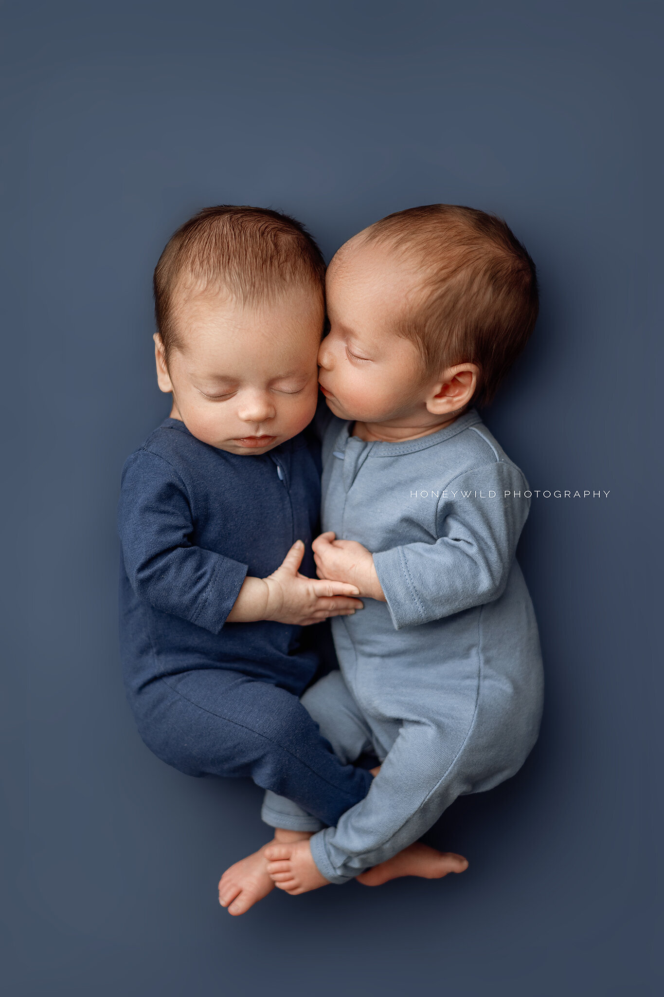 Twin newborns in blue pajamas cuddled together on a navy backdrop during a studio newborn session in Grand Rapids, MI by Honeywild Photography.