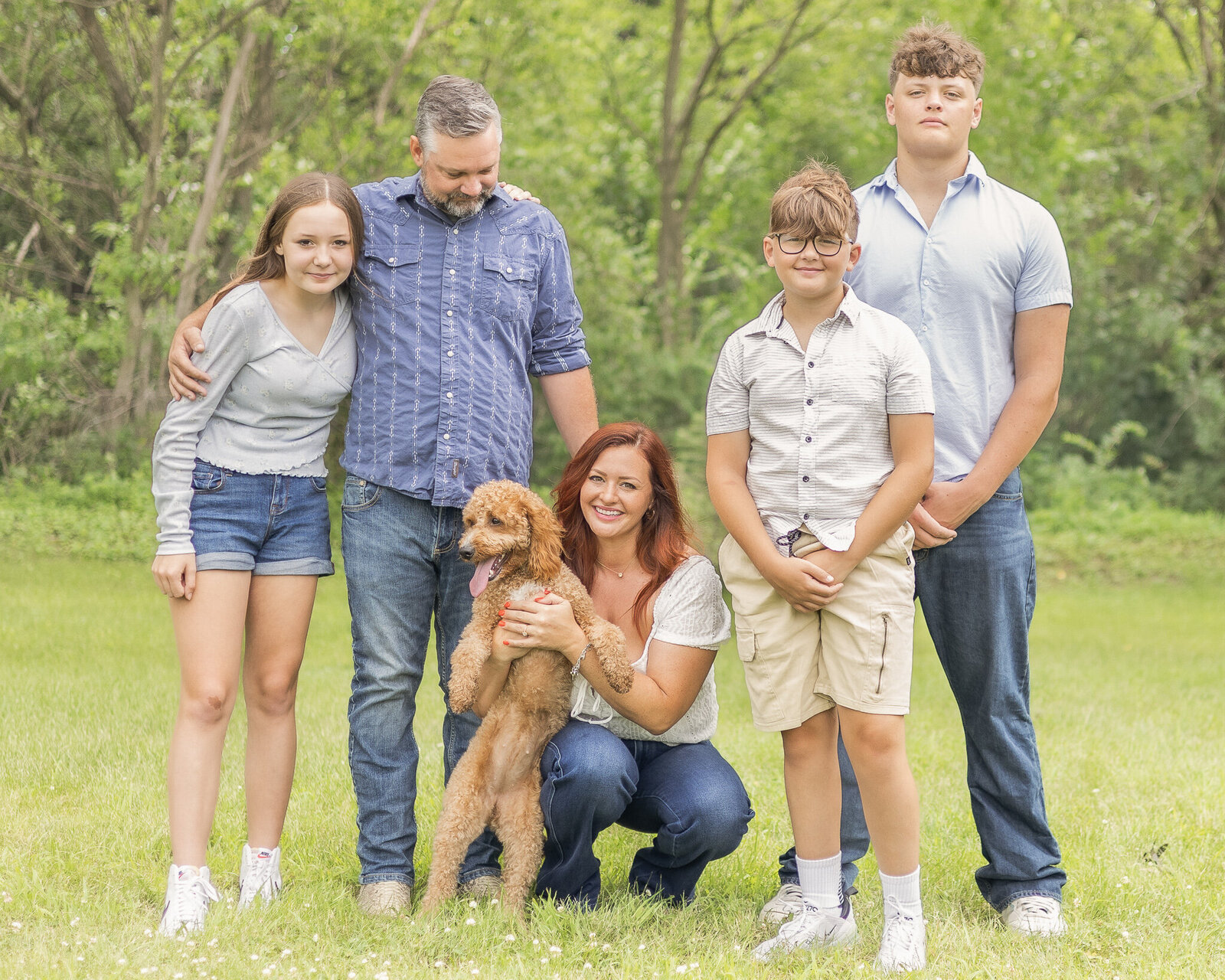 A family of 5 and their brown dog standing in the grass.  Dad has brown gray hair, wearing a blue and white striped shirt, blue jeans, and is looking down at his wife.  Daughter is wearing blue jean shorts and a light blue shirt, with brown hair, smiling at the camera.  Mom is knelt down with their brown dog, with red hair, wearing blue jeans, a white shirt, and smiling at the camera.  Younger son has brown hair, wearing a light tan shirt and shorts, smiling at the camera.  The oldest son has  brown hair wearing a light blue shirt and blue jeans, looking at the camera with a straight face.  The brown dog is standing on it's back feet with tongue hanging out and looking off camera