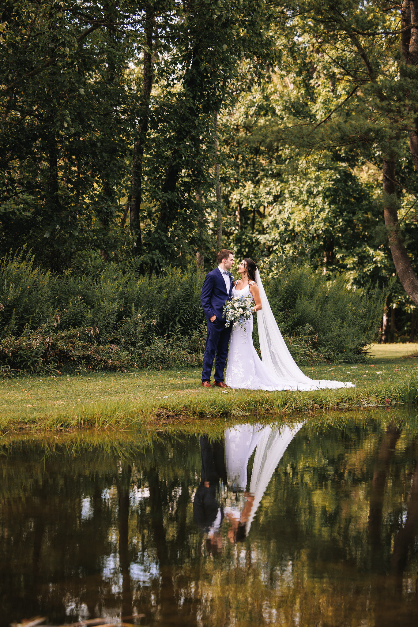 A bride and groom gaze at each other in front of a pond with a lush green forest in the background. Their reflection in the water below