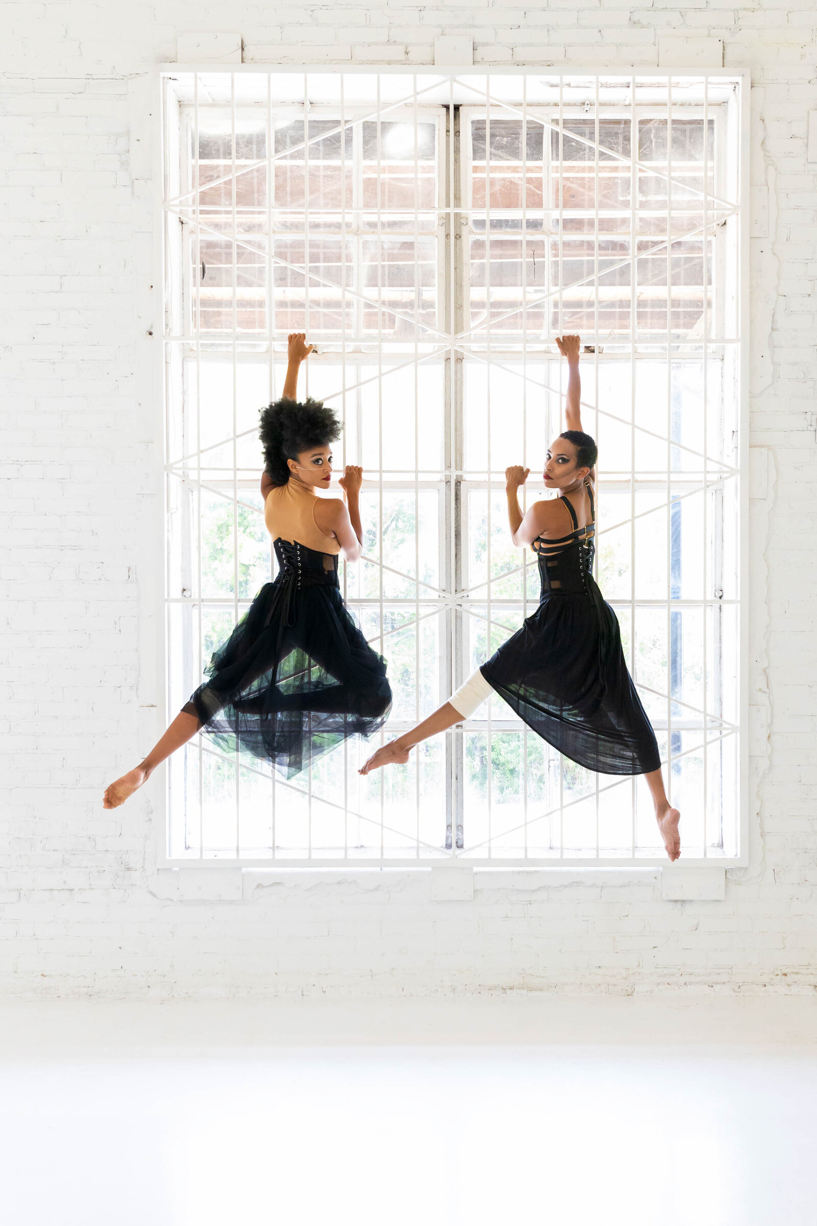 two black women dancers in sheer skirts and bustiers hanging on to window