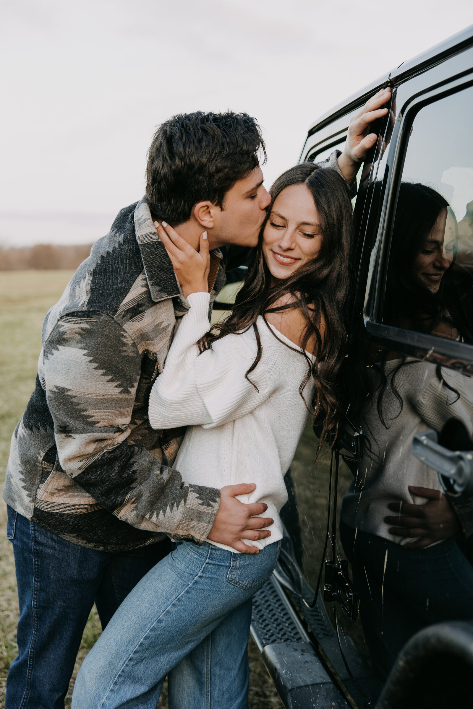 A couple leans against a Jeep; the man kisses her head while she smiles warmly at the camera, bathed in sunlight.