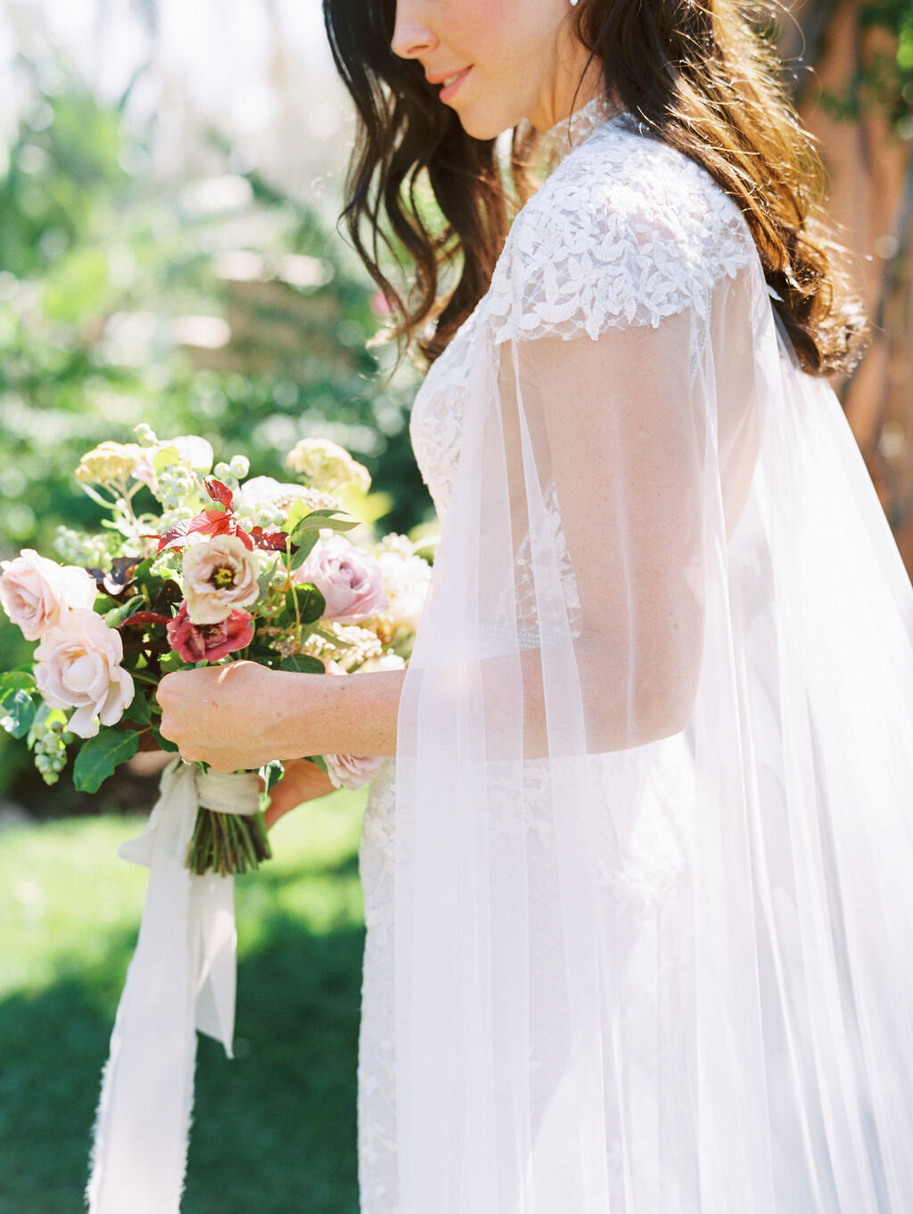 Bride holding bouquet wearing a lace gown by Berta with a tulle cape
