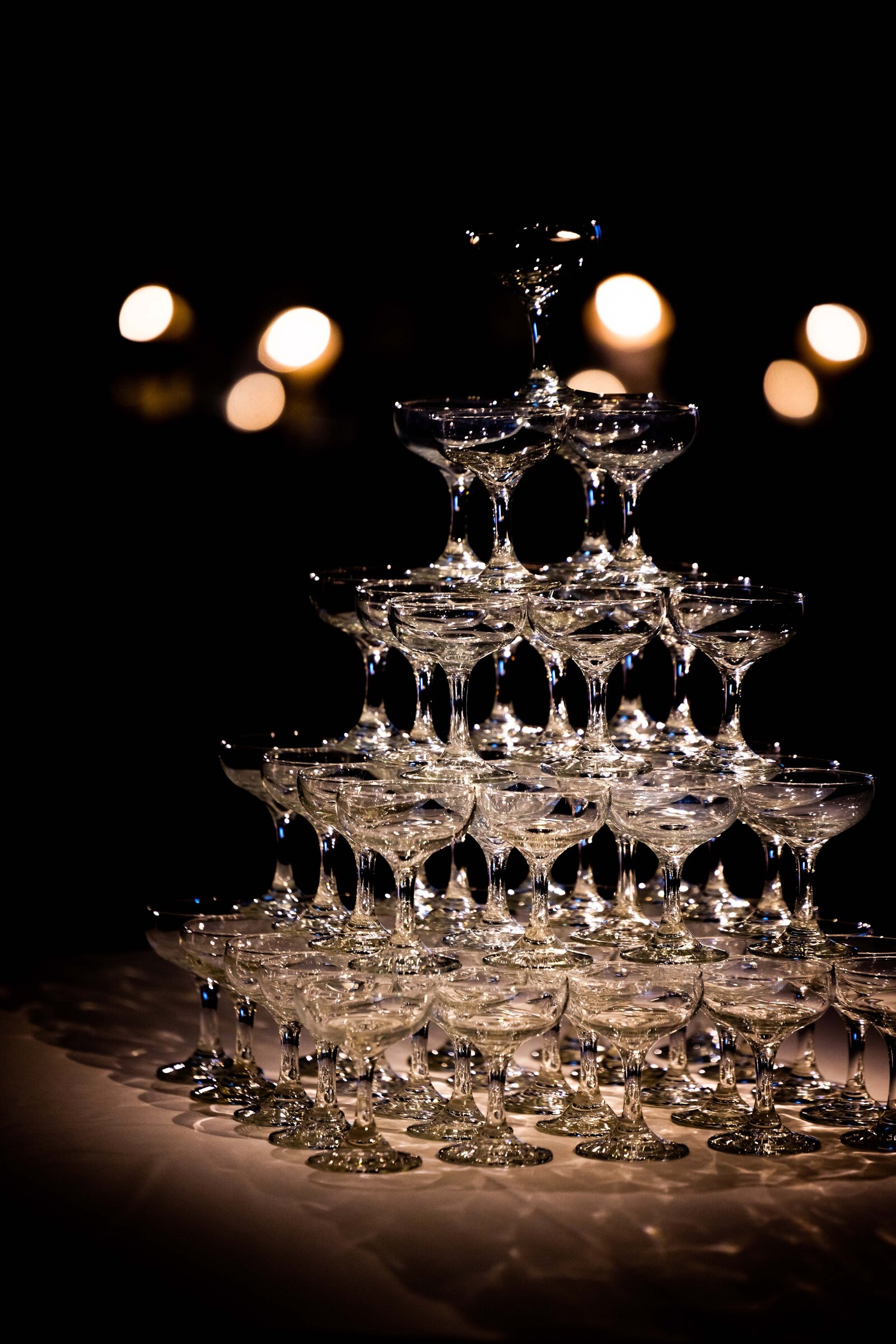 Step into a scene of dramatic elegance with this moody, backlit photograph of an extravagant champagne tower. The soft backlighting casts a mysterious glow, enhancing the allure and sophistication of the towering glasses filled with bubbling champagne. This captivating image is perfect for those seeking to add a touch of opulence and theatrical flair to their event, offering a visually stunning centerpiece that embodies celebration and luxury.