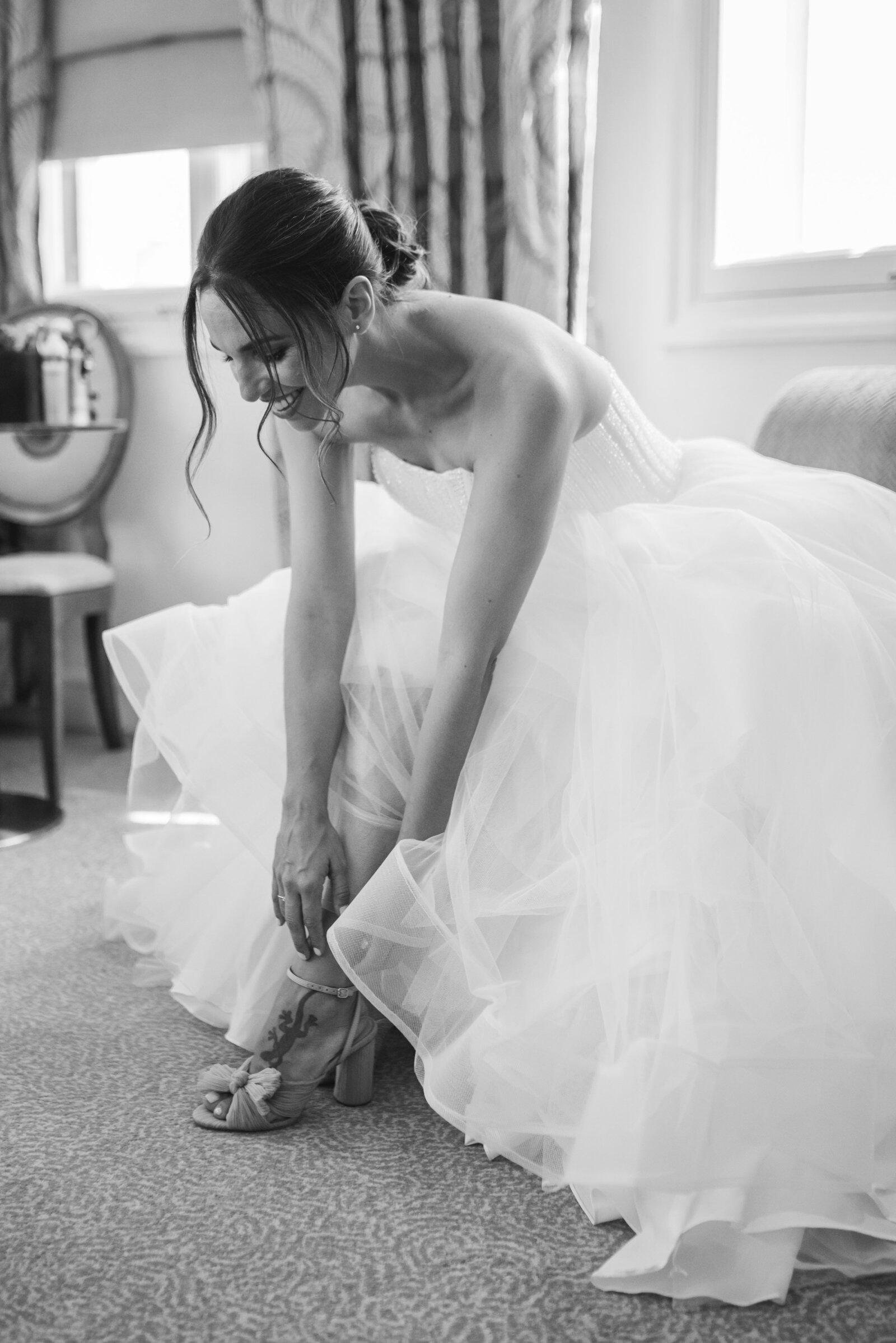 Black and white image of a bride putting on her wedding shoes. The bride is looking down and smiling and is in a ballgown style dress.