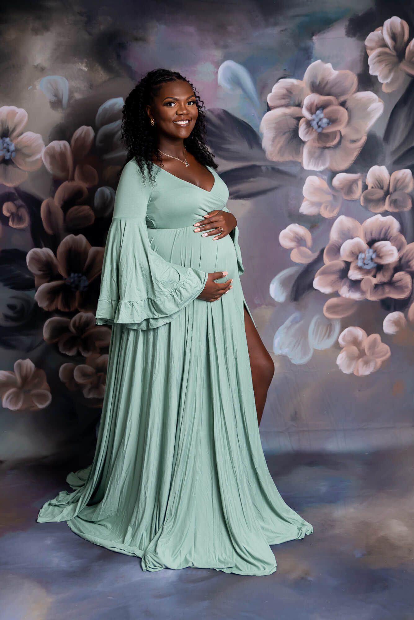 A woman in a sage green gown poses in front of a painted floral backdrop, smiling as she cradles her belly.