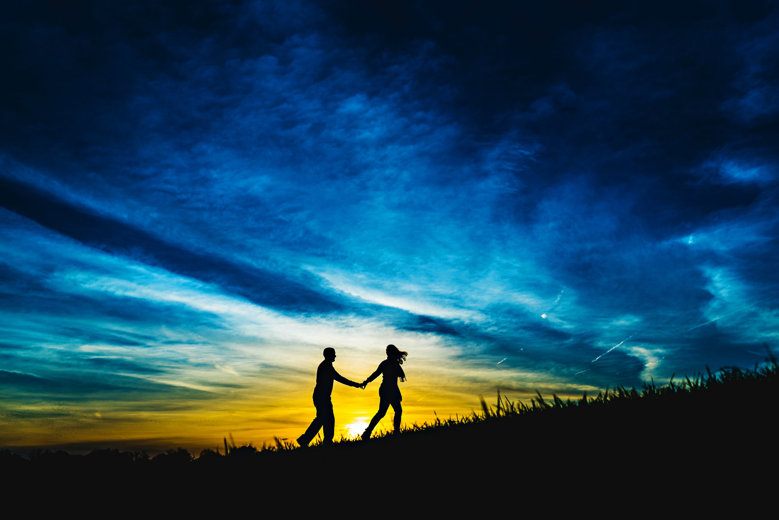An engaged couple walk up a hill in the setting sun.