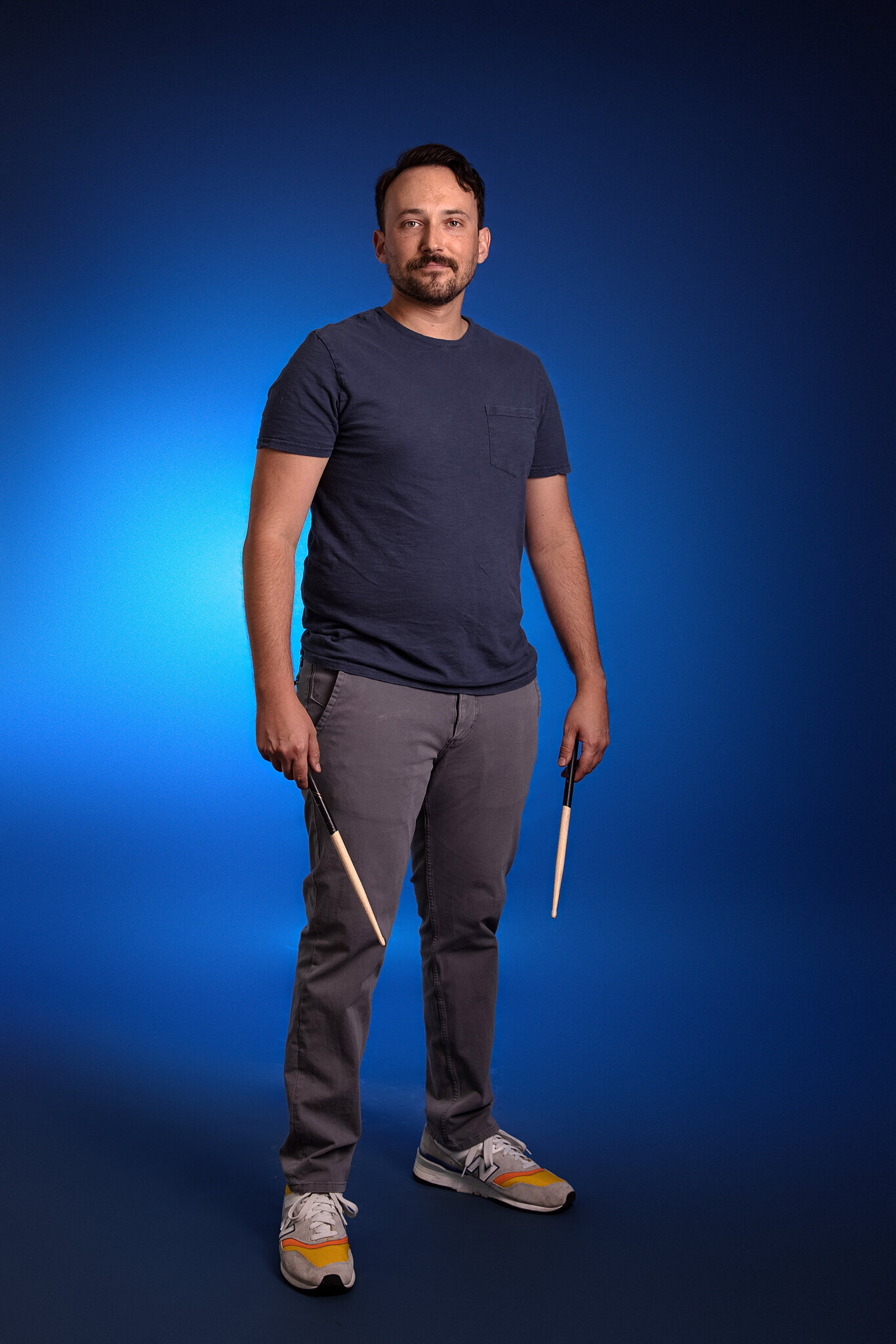 A man with short dark hair, a beard, and a mustache stands against a blue background, wearing a navy t-shirt, gray pants, and sneakers, holding drumsticks—a great example of band photography Jacksonville style.