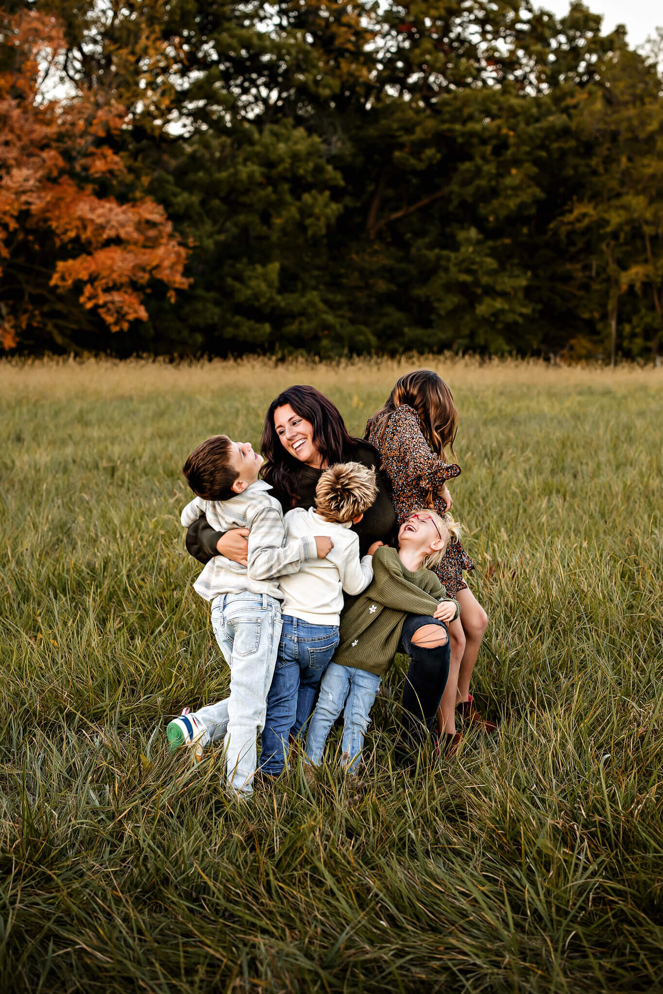 In a grassy field, a woman joyfully embraces four children, framed against a backdrop of green trees and orange foliage. This heartwarming outdoor portrait captures a moment of happiness and connection, showcasing the talent of a skilled Bloomington family photographer.
