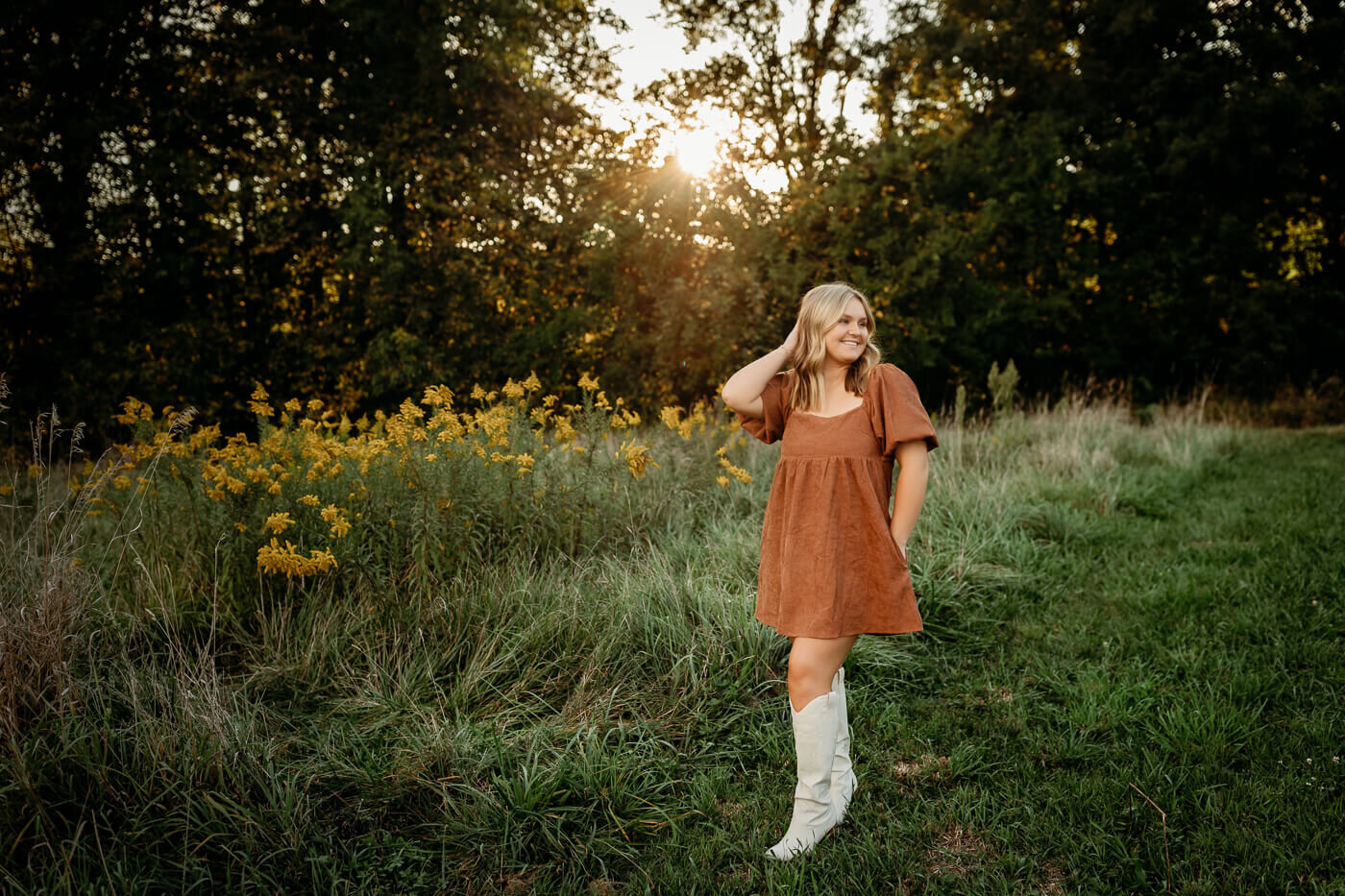 A person with long blonde hair stands in a grassy field at sunset. They are wearing a brown dress and white boots, surrounded by wildflowers and trees in the background.