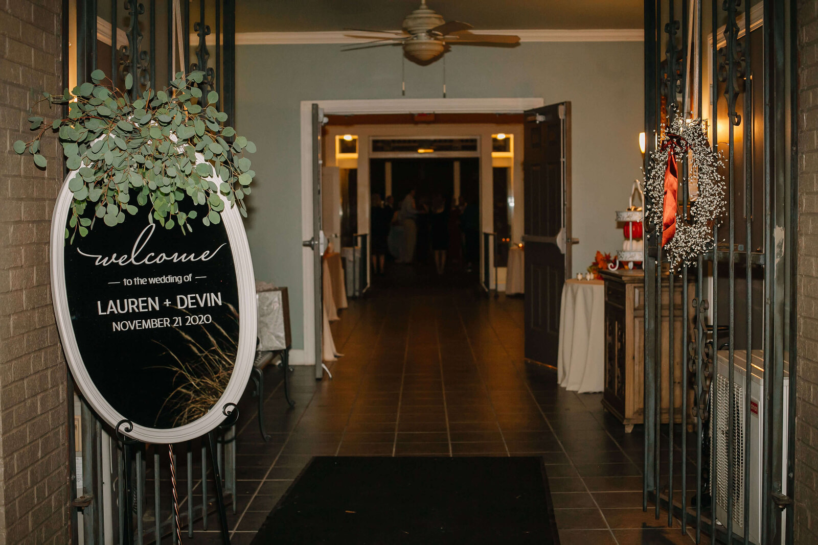 A wedding entrance with a welcome sign for Lauren and Devin. The hallway is warmly lit, decorated with greenery and autumn wreaths, leading to a room with people in the distance.