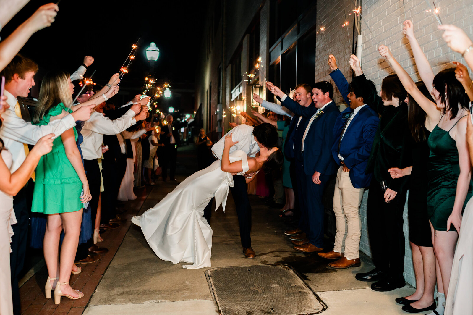 A bride and groom share a kiss as the groom dips the bride, surrounded by cheering guests holding sparklers and celebrating their wedding in a nighttime outdoor exit.
