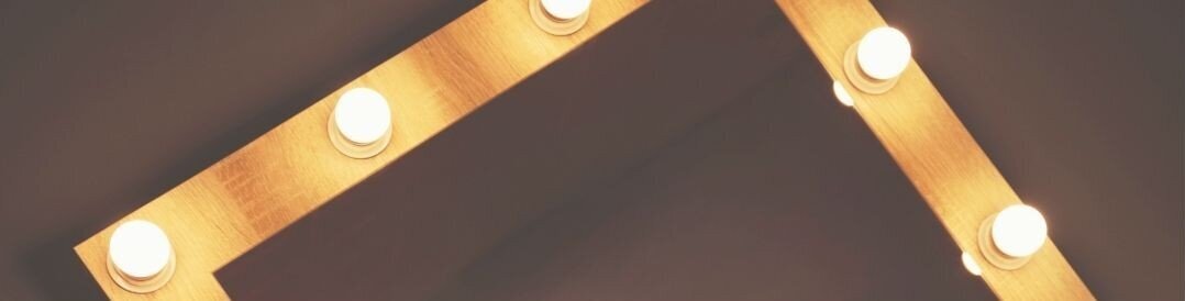 Close-up of a vintage-style vanity mirror with glowing Hollywood bulbs, used as a background banner for Las Vegas bridal hairstyling services.