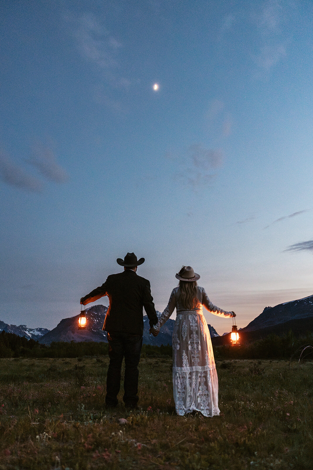 A couple holds lanterns and looks up at the night sky under the moon in Glacier National Park, surrounded by mountains and open fields, captured by Sydney Breann Photography during their adventure elopement.