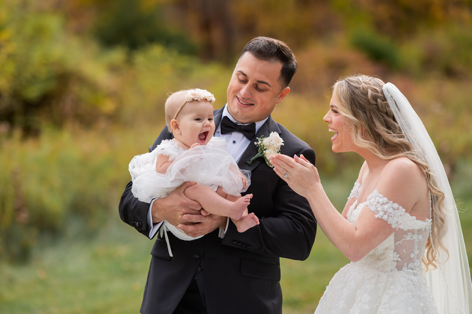A bride and groom stand outdoors in formal attire, holding and smiling at a baby in a white dress.