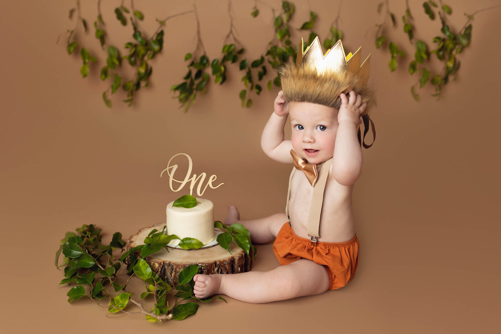 A joyful baby with blond hair and blue eyes sits on a beige backdrop with hanging greenery, wearing an orange diaper cover, beige suspenders, and a golden crown with a faux fur base. The baby smiles while holding the crown on their head. In front of them is a rustic cake on a wooden slab, decorated with green leaves and topped with a "One" cake topper, celebrating their first birthday.