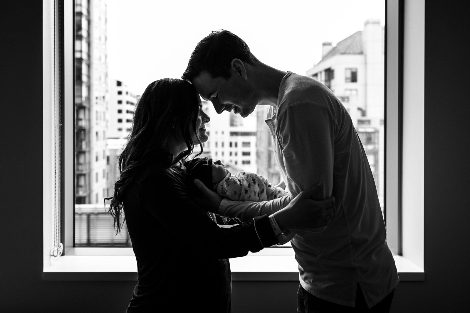 parents standing in front of window holding swaddled newborn and touching foreheads in San Francisco hospital